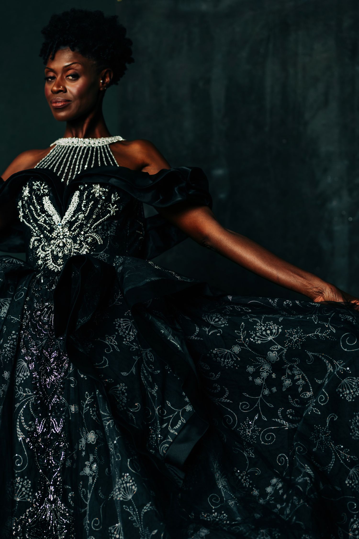 Elegant woman in black embroidered ballgown with pearl necklace posing gracefully against dark backdrop.