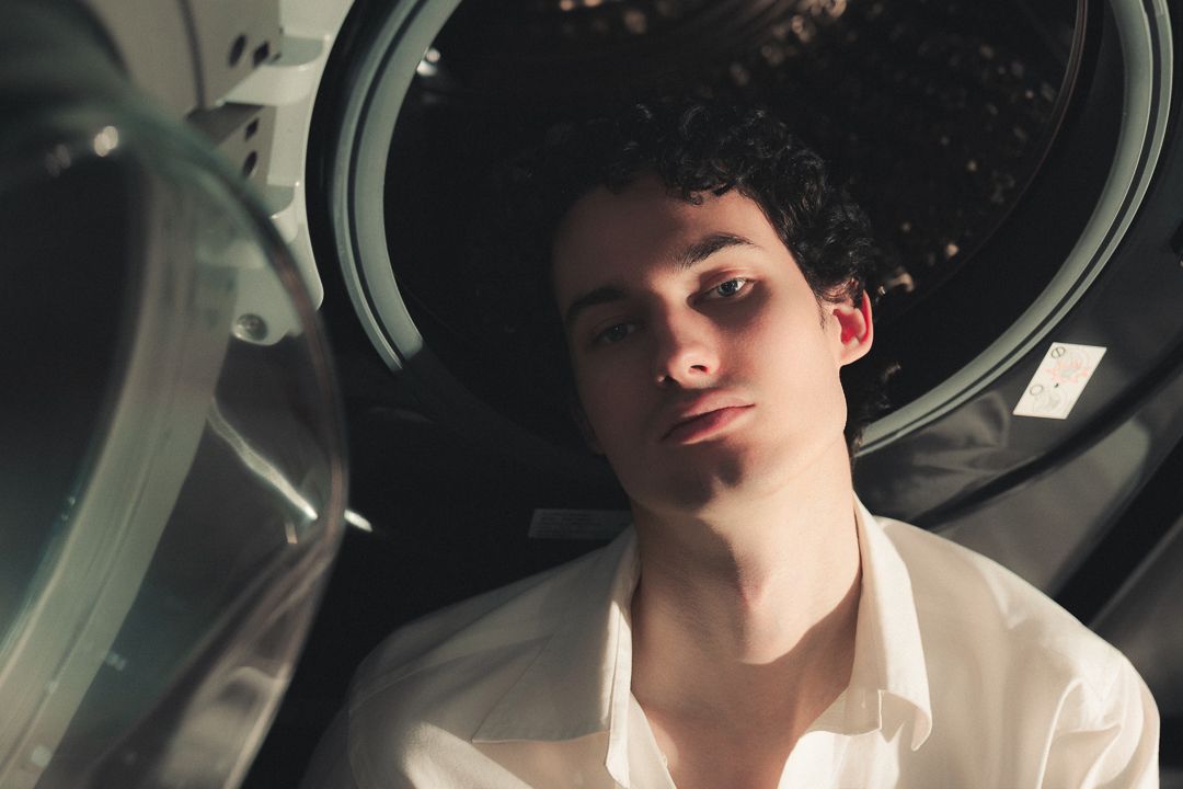 Young man with curly hair in white shirt lying near a washing machine drum, artistic portrait photography.