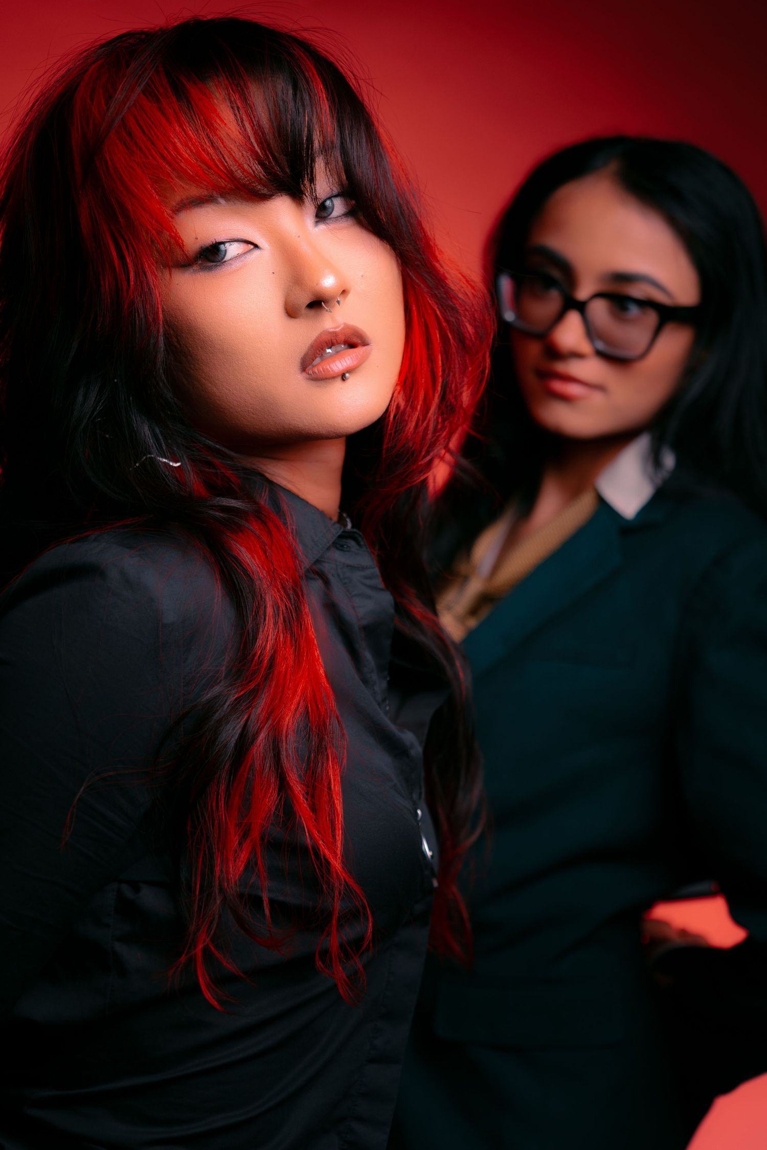 Two young women with dark hair pose against a red background, one with red highlights wearing a black shirt.
