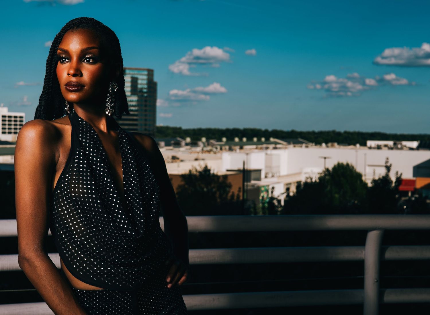 Confident young Black woman in a sparkly halter top posing on a rooftop with city skyline behind her.