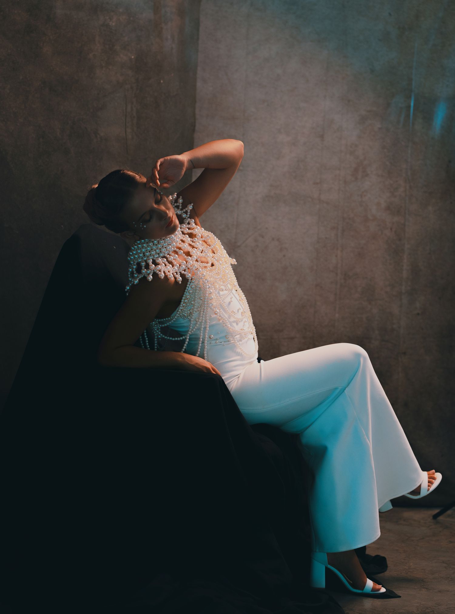 Pregnant woman in white lace top and wide-leg pants sits gracefully in dark studio chair with dramatic lighting.