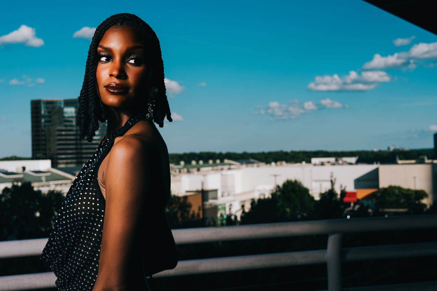 Young Black woman in polka dot dress poses on urban rooftop balcony with city skyline and blue sky.