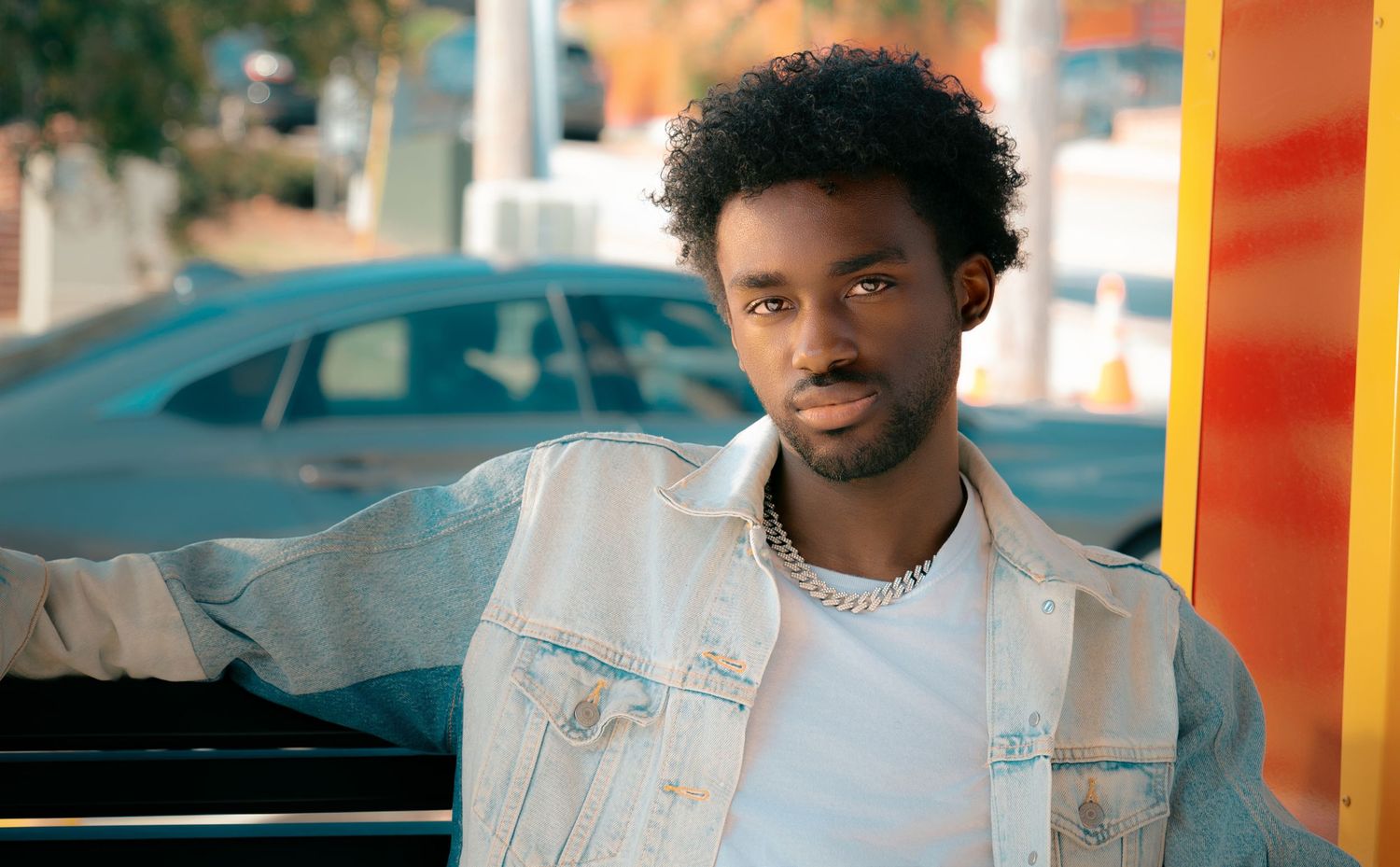 Young man with curly hair wearing a light denim jacket and chain necklace, posing outdoors near a colorful urban setting.