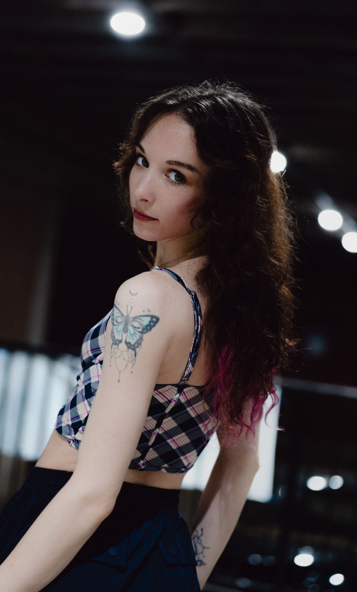 Young woman with curly dark hair, butterfly tattoo on shoulder, wearing plaid crop top, posing in dimly lit studio.