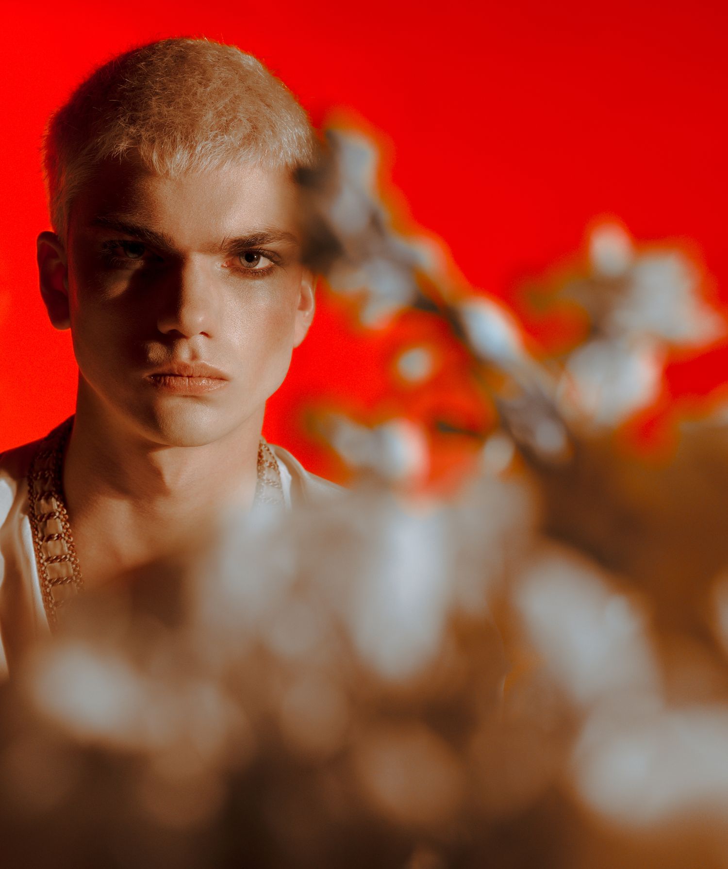 Young man with blonde hair poses against a vibrant red background with blurred white flowers in the foreground.