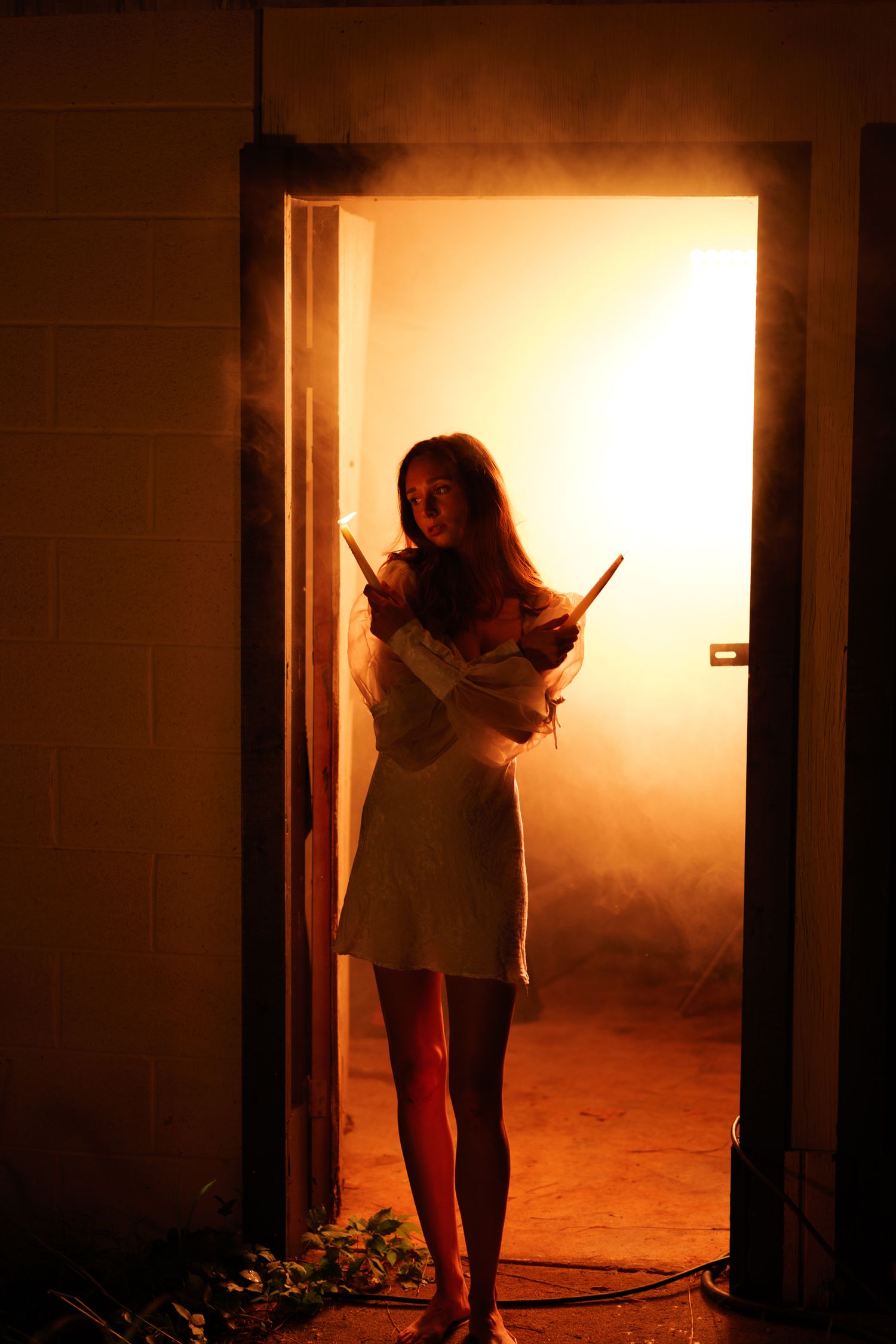 Woman in white dress holding sticks stands in smoky doorway with dramatic orange fiery glow behind her.