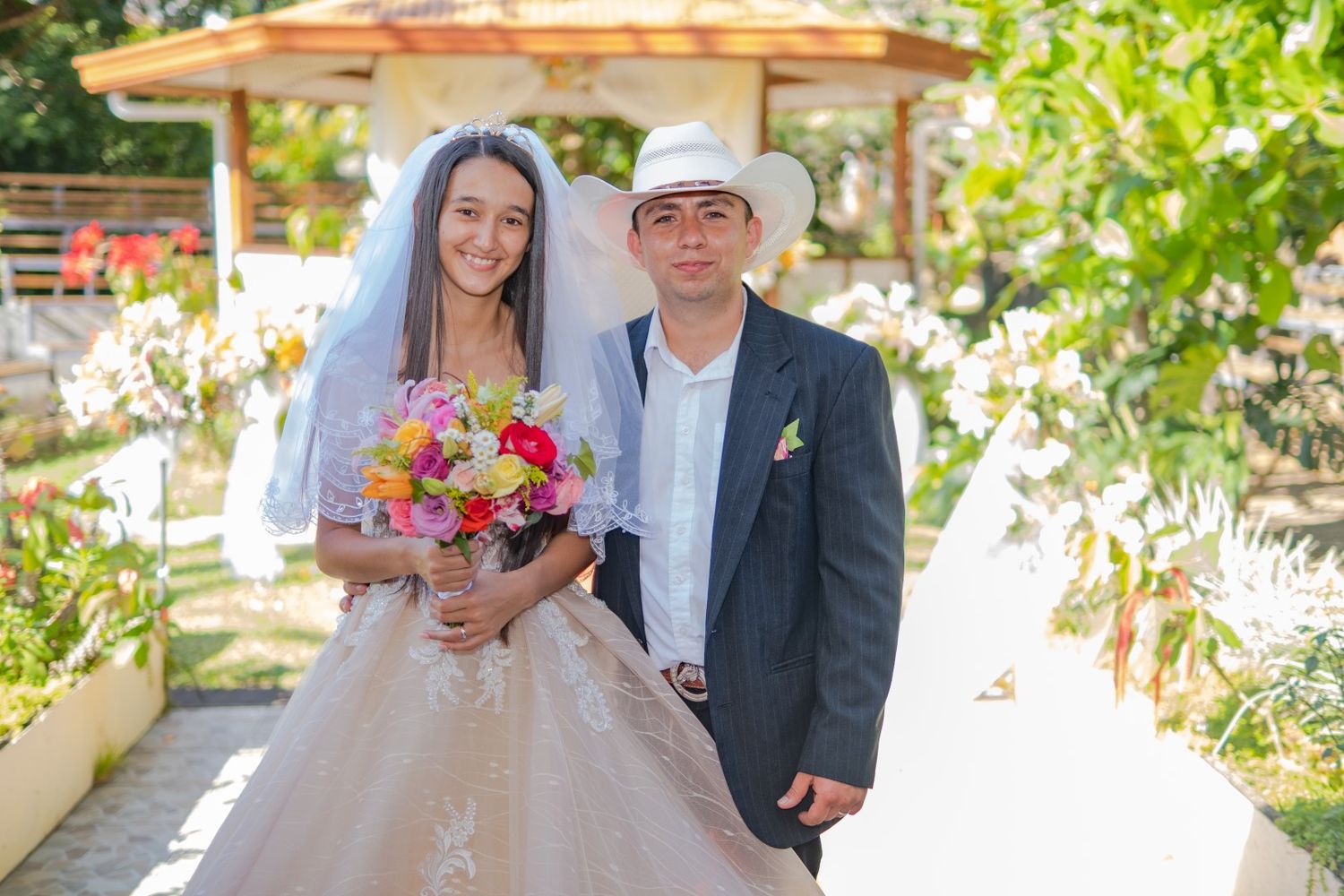 Wedding couple with colorful bouquet posing under garden gazebo in formal attire.