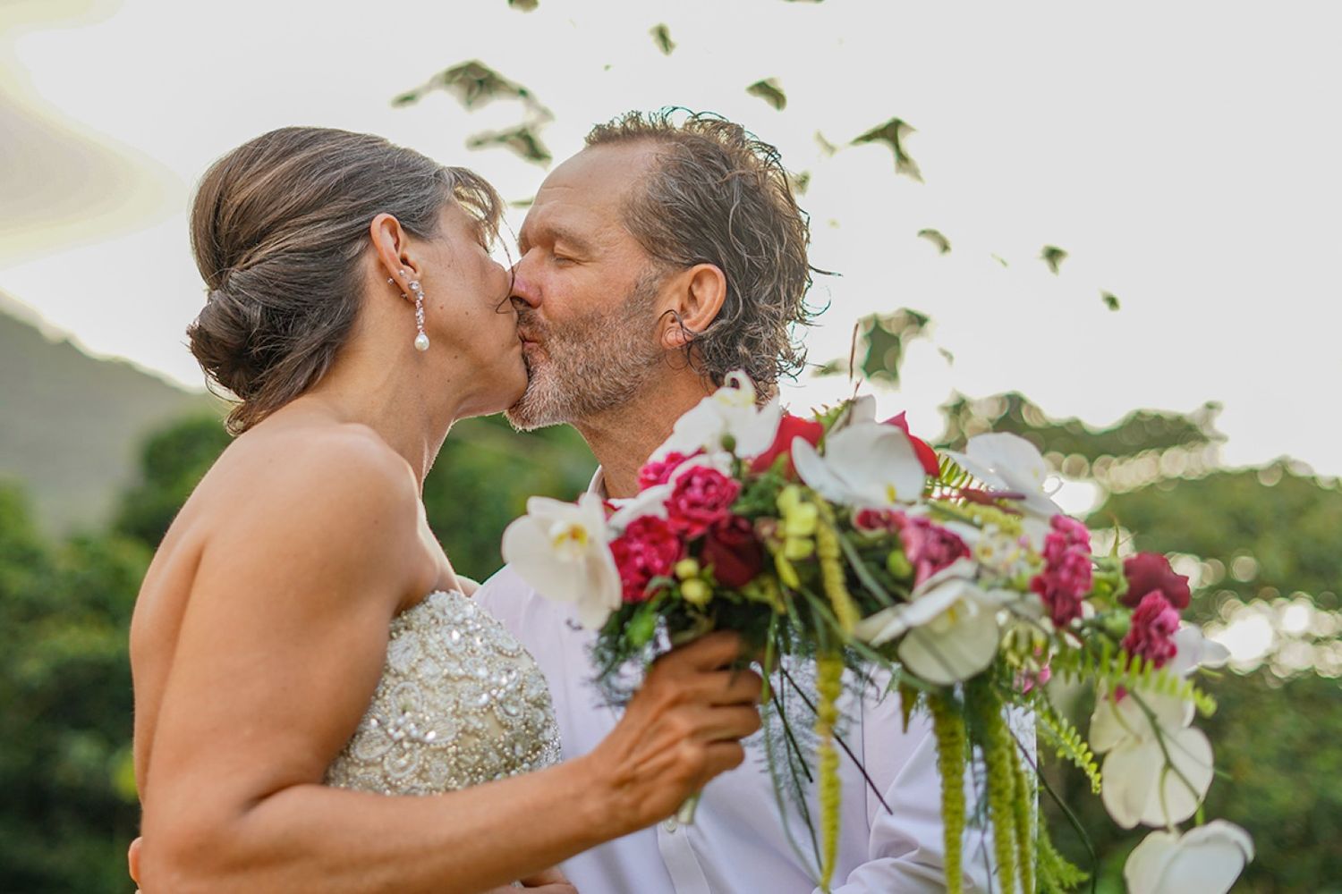 Romantic outdoor wedding photo with couple holding vibrant red bouquet among greenery.