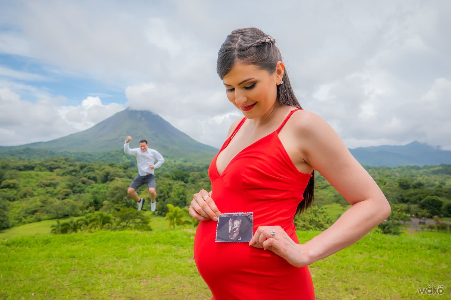 Pregnant woman in red dress poses while partner jumps playfully in mountain background.