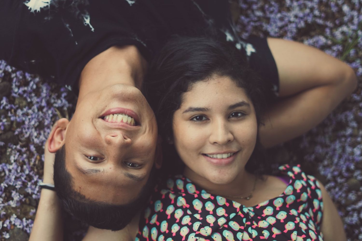 A couple lying on grass together smiling at the camera in a romantic outdoor setting.