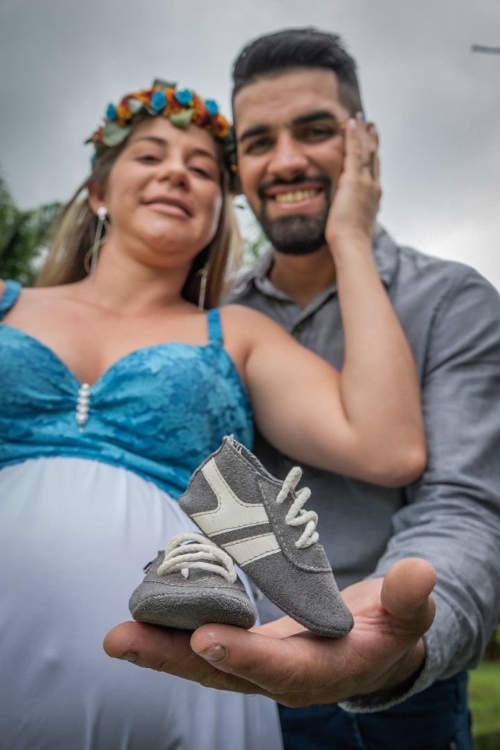 Expecting couple smiles while holding small gray baby shoes outdoors.