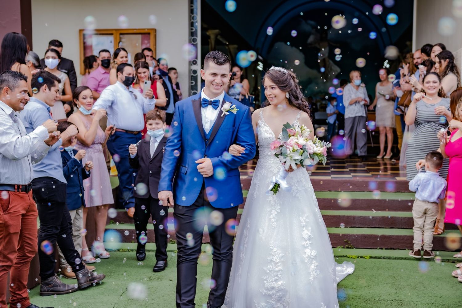 Wedding couple exits ceremony while guests blow bubbles in celebration.