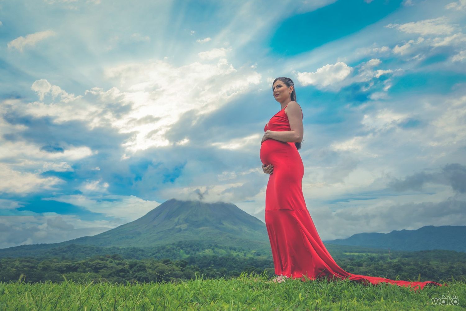 Expectant mother in flowing red dress poses against mountain landscape during sunset.