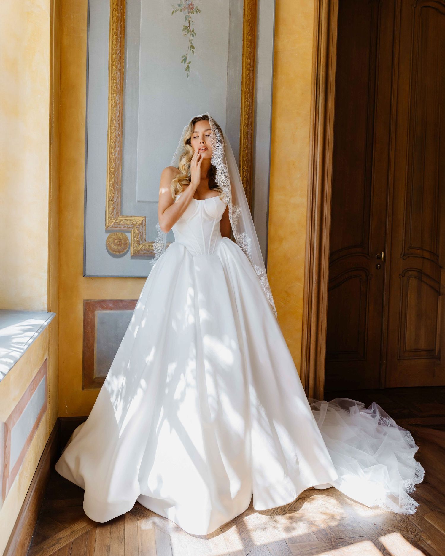 Bride in a white ball gown with veil stands in sunlit ornate room.