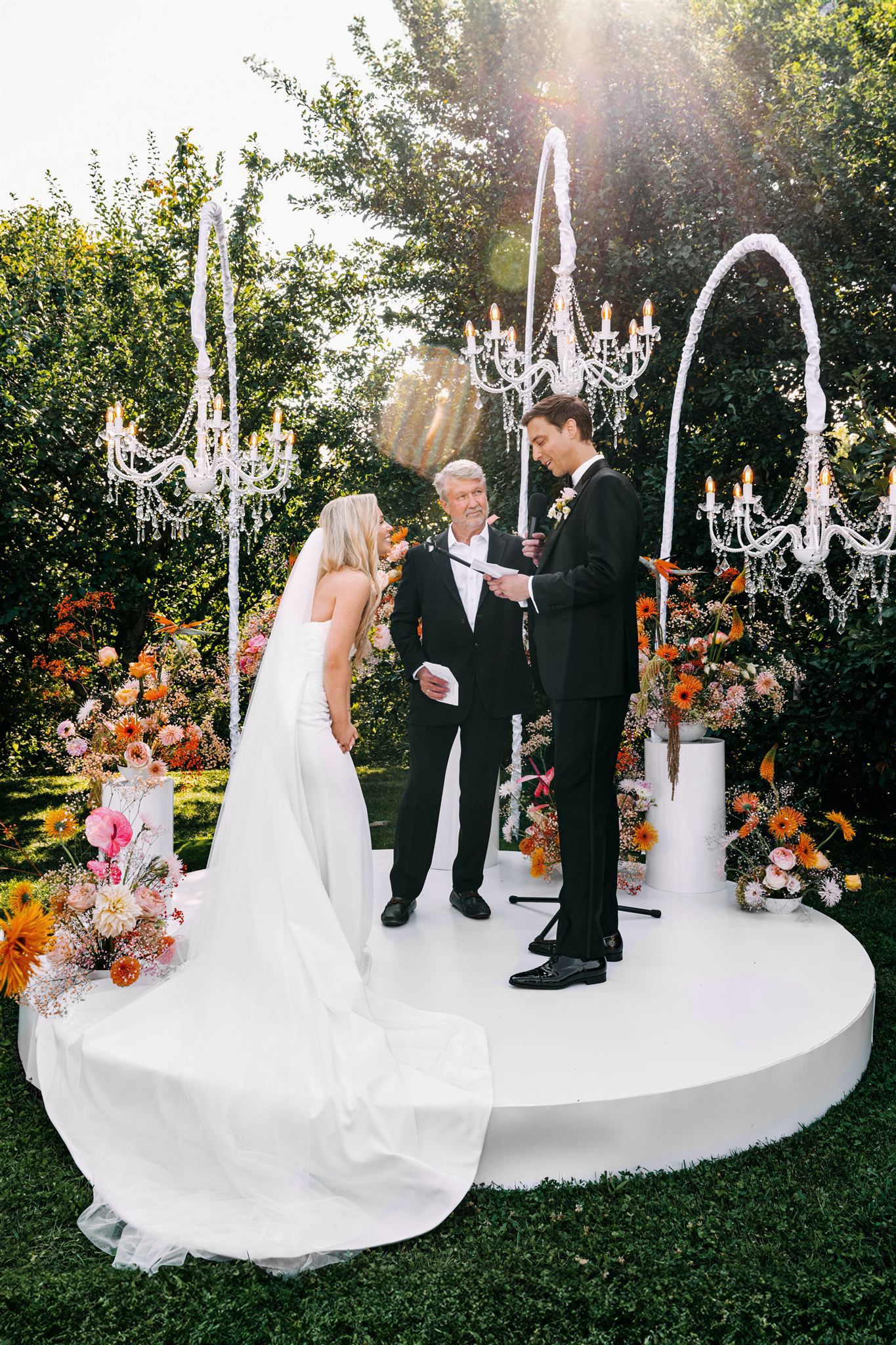 Outdoor wedding ceremony on circular white platform with hanging chandeliers and floral decor.
