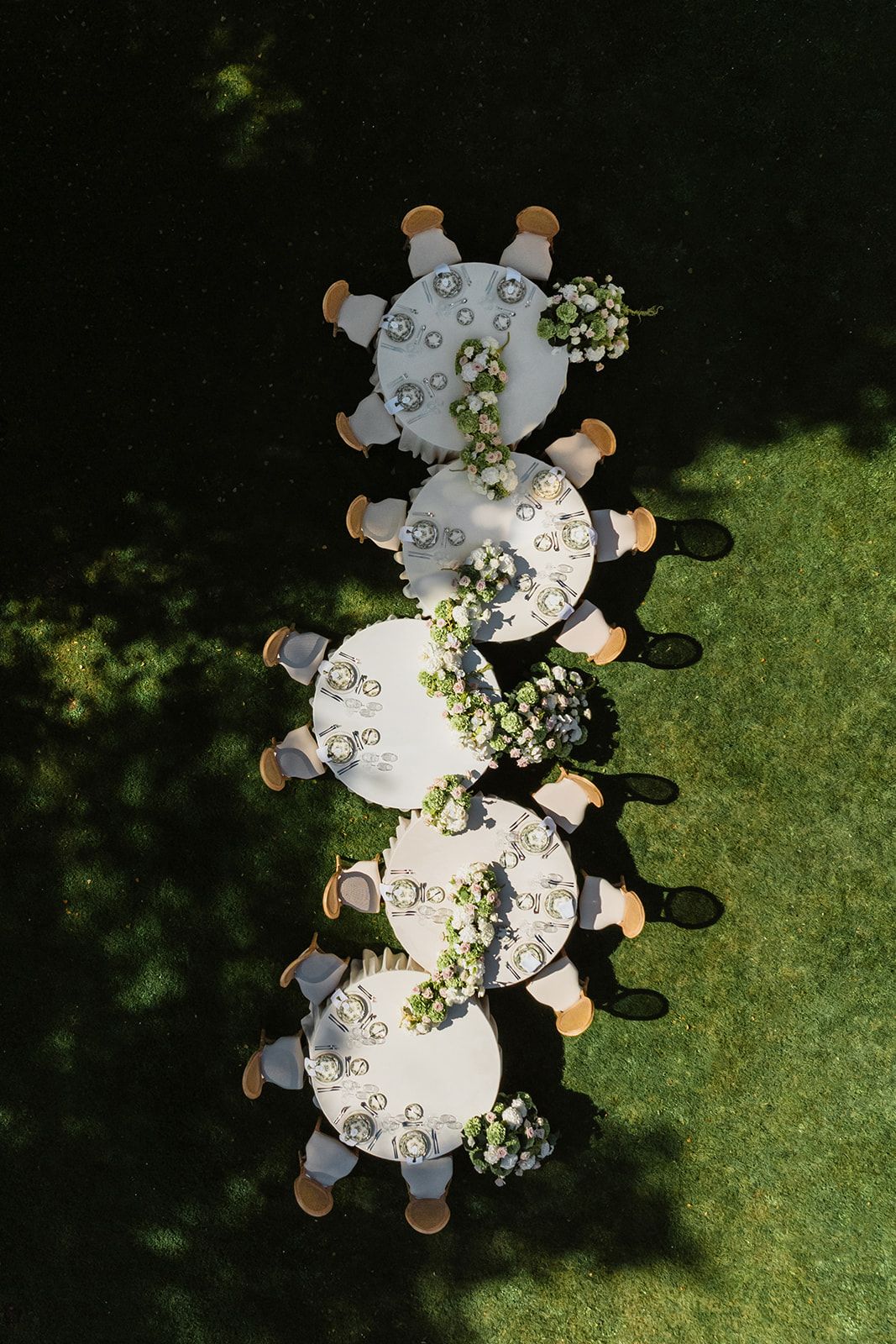 Aerial view of circular wedding reception tables with white linens and floral centerpieces on grass.