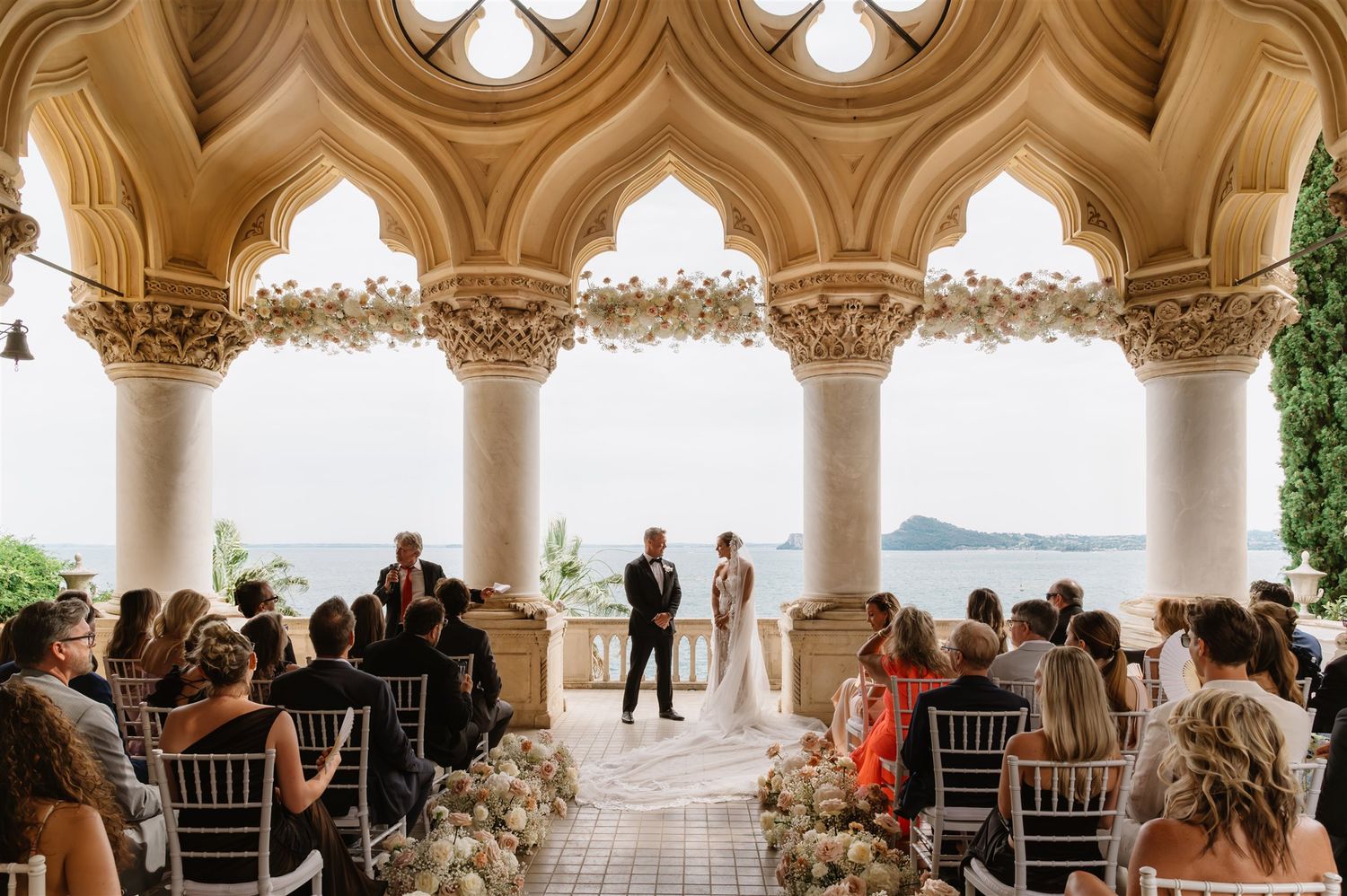 Wedding ceremony setup under elegant stone arches with ocean views and white chairs.