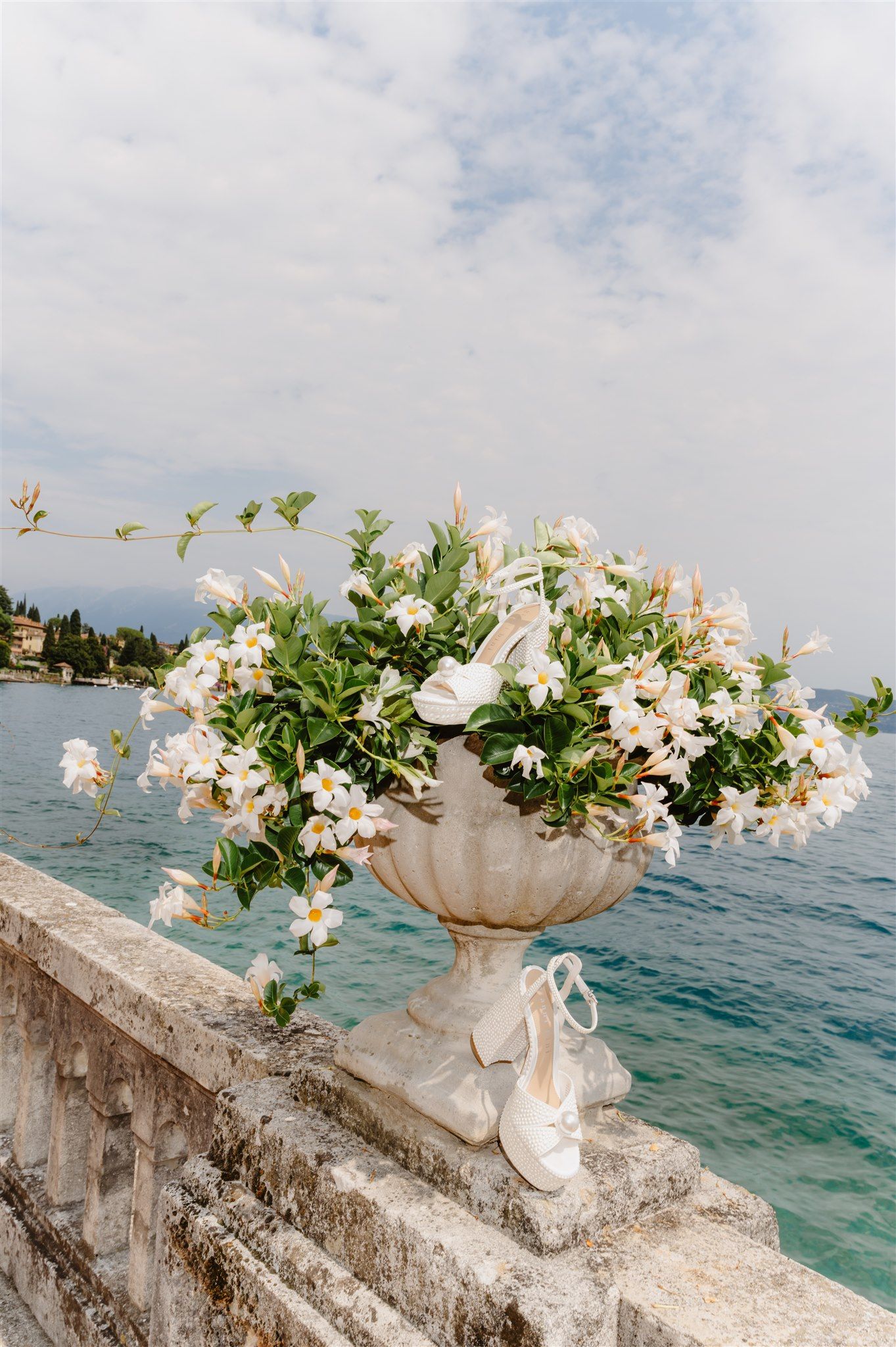 White floral arrangements in stone vases line a waterfront balustrade overlooking Lake Garda.