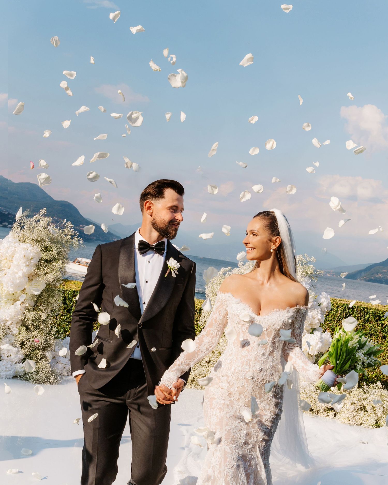 White flower petals float through the air around a bride and groom on their wedding day with mountains in background. Lago Maggiore.
