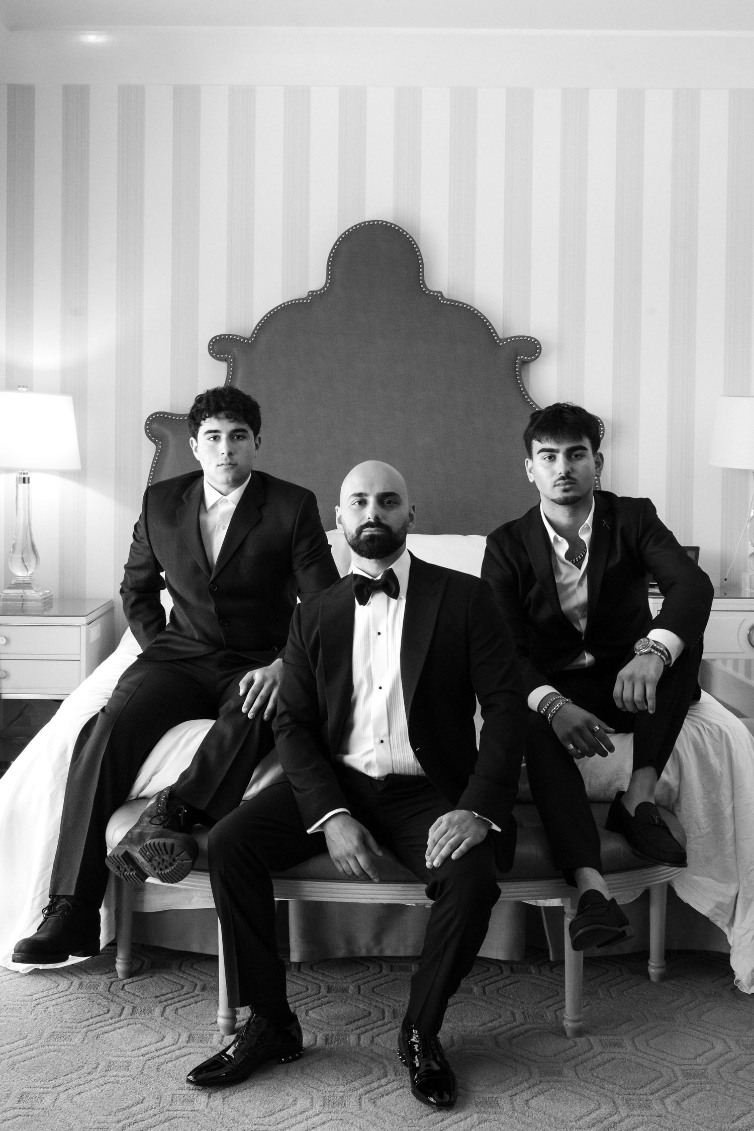 Three men in formal black tuxedos pose elegantly on a bed in a black and white portrait photo.