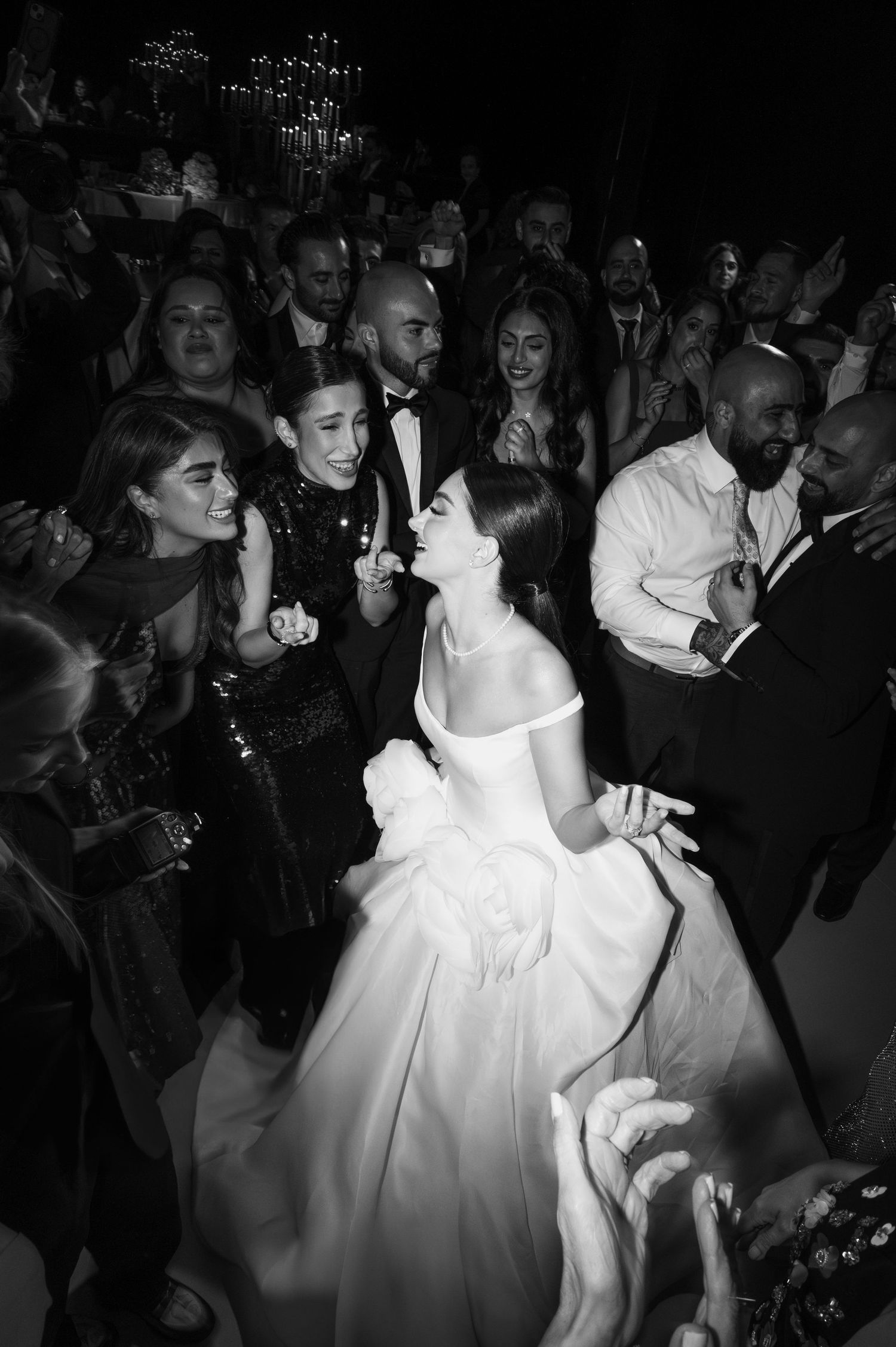 Black and white photo of a bride in an off-shoulder gown dancing joyfully surrounded by wedding guests.