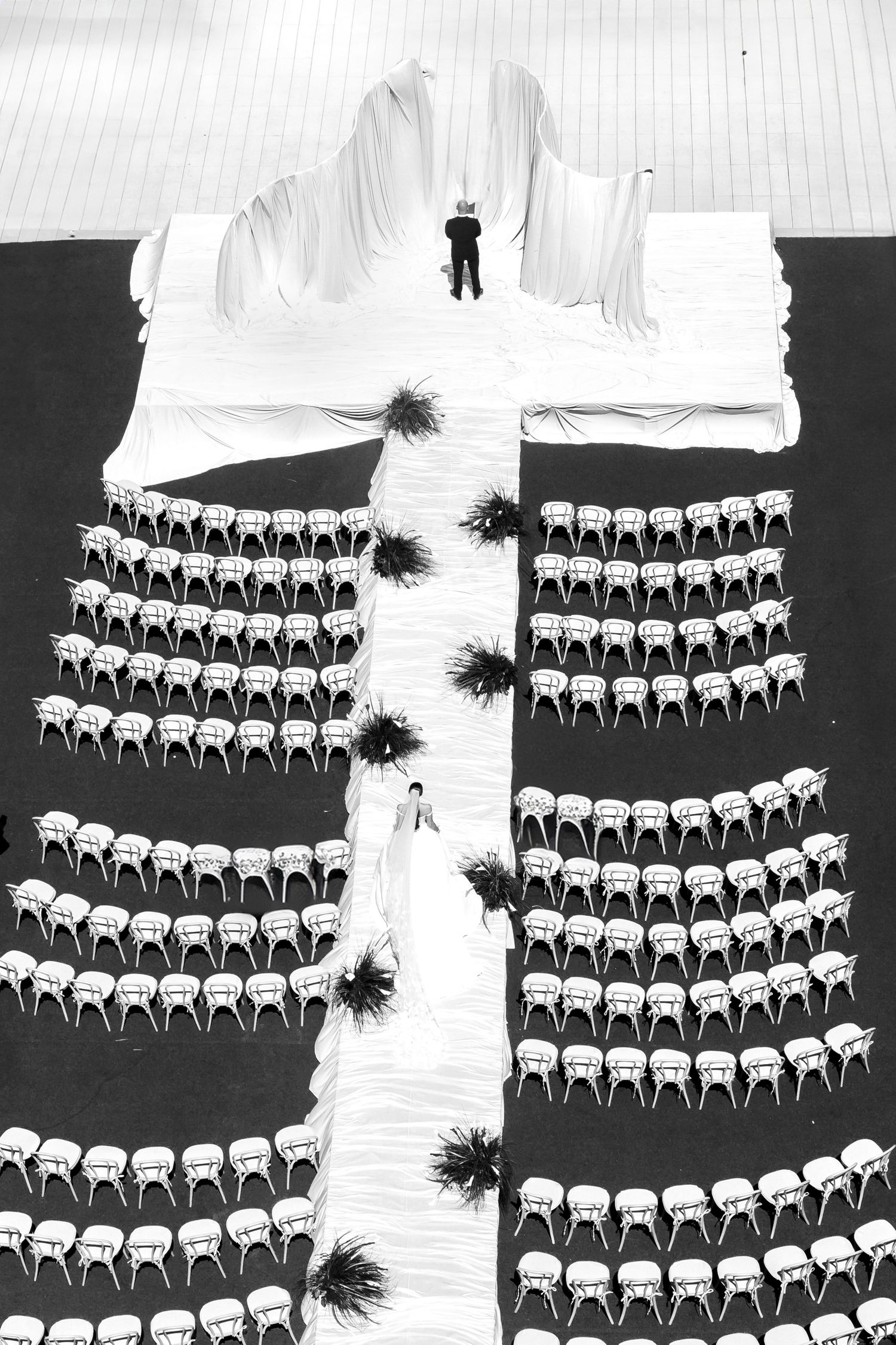 Aerial black and white view of wedding ceremony setup with rows of chairs forming a cross shape on dark ground.
