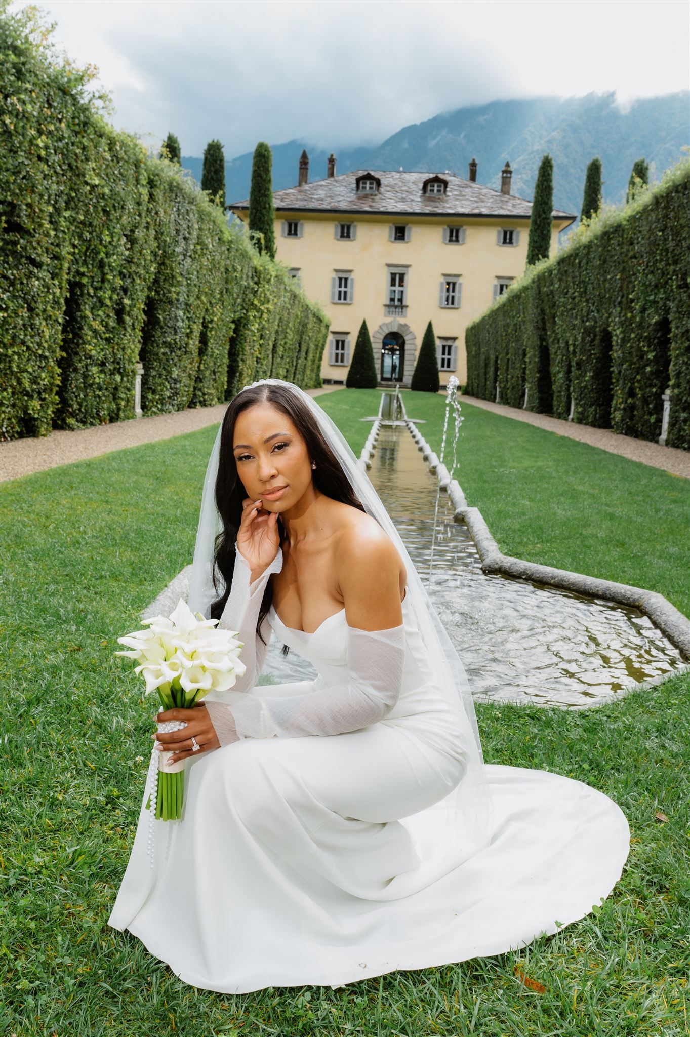 Bride in flowing white gown poses gracefully on manicured lawn with Italian villa backdrop.