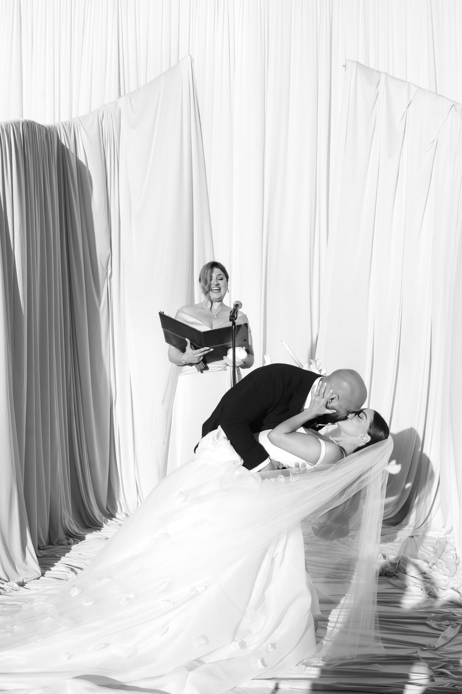 Black and white photo of groom dipping bride during wedding ceremony with officiant reading in background.