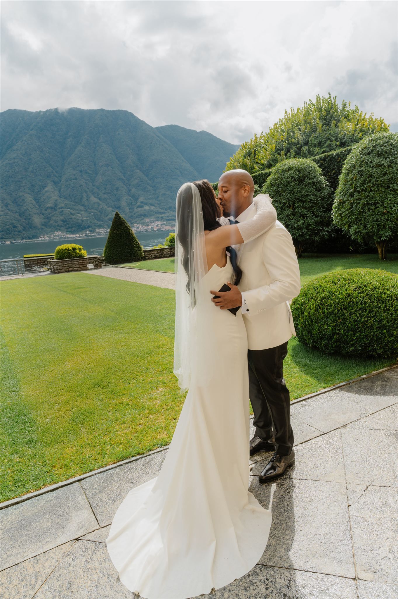 A romantic wedding couple embraces in a garden with mountain views and manicured hedges.