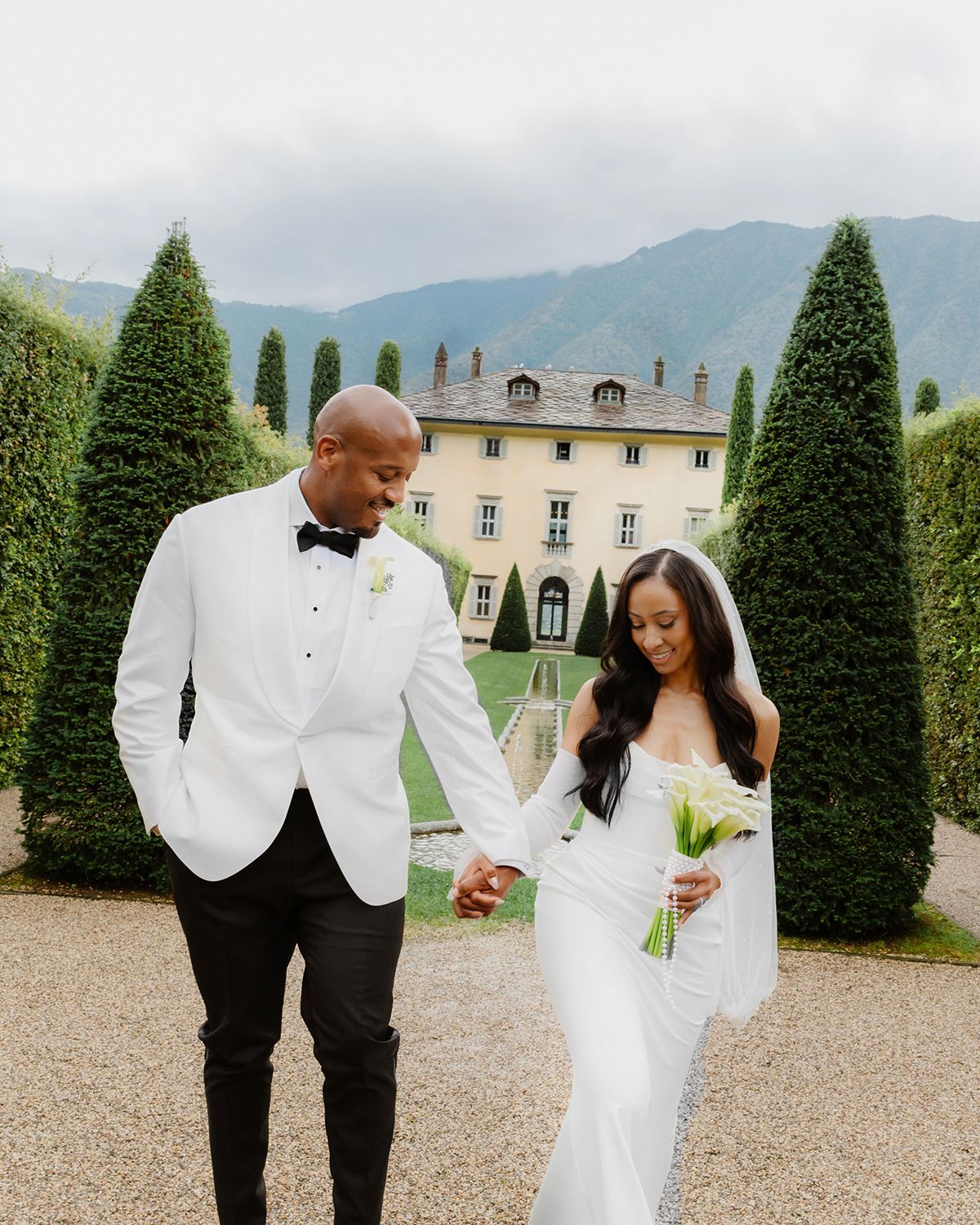 A couple shares a joyful moment together on a gravel path surrounded by manicured topiary trees and a stately mansion.