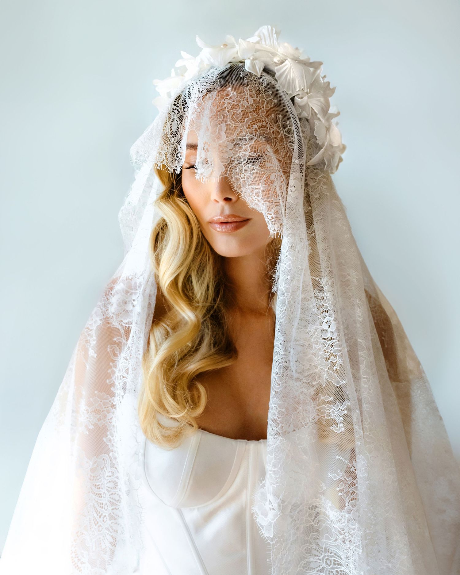 Bride wearing an elegant lace mantilla veil with floral crown accents over flowing blonde hair and white gown.