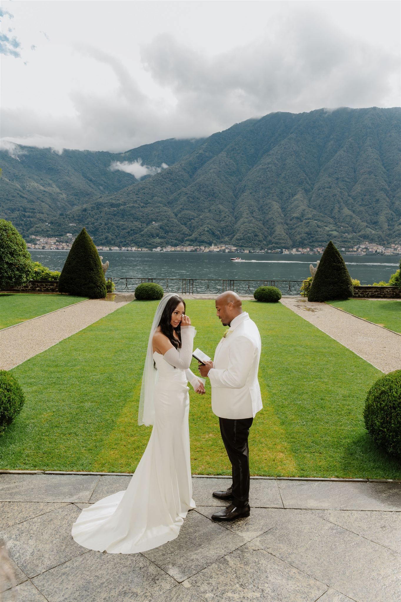 A couple in white wedding attire poses against a backdrop of mountains and manicured gardens.