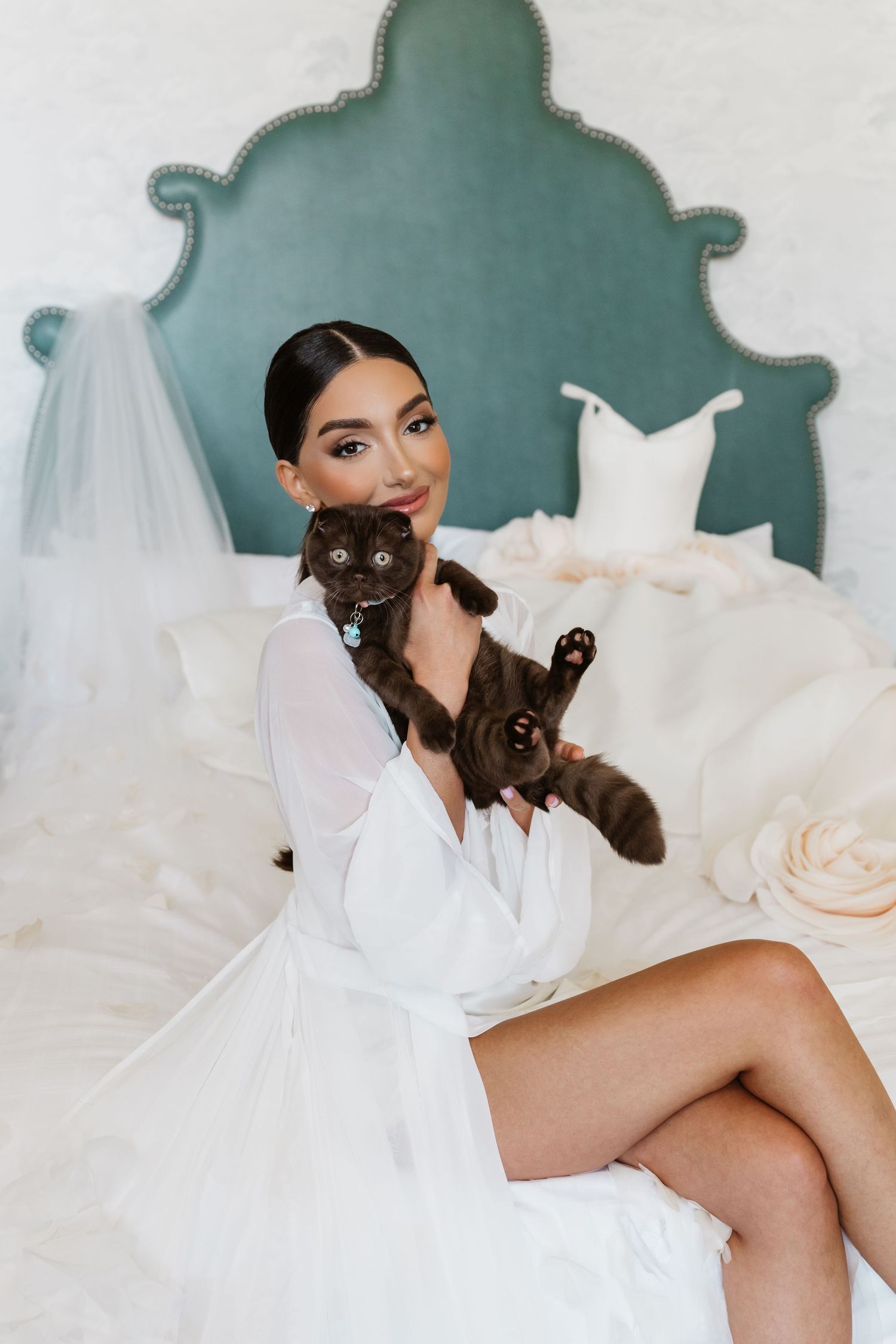 Bride in white robe holds dark cat on bed with wedding dress and veil in background.
