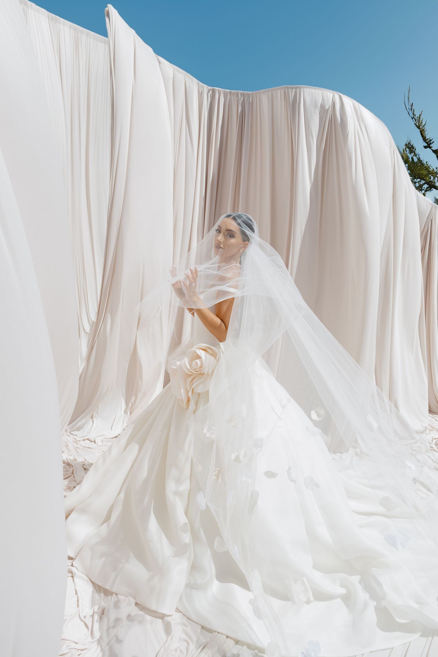 Bride in a stunning white ball gown with floral details and long veil poses among flowing white drapes under blue sky.