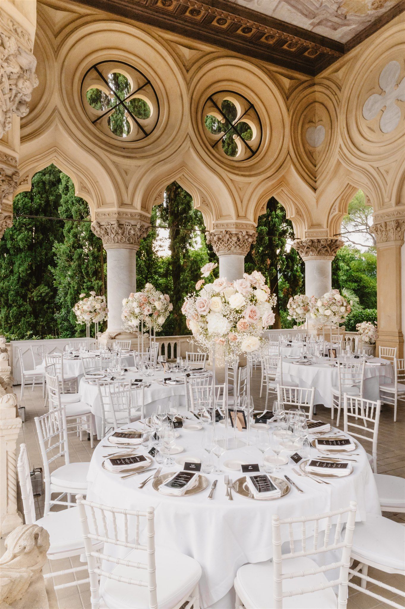 Luxurious white wedding reception setup in an ornate colonnade with floral centerpieces. Isola del garda.