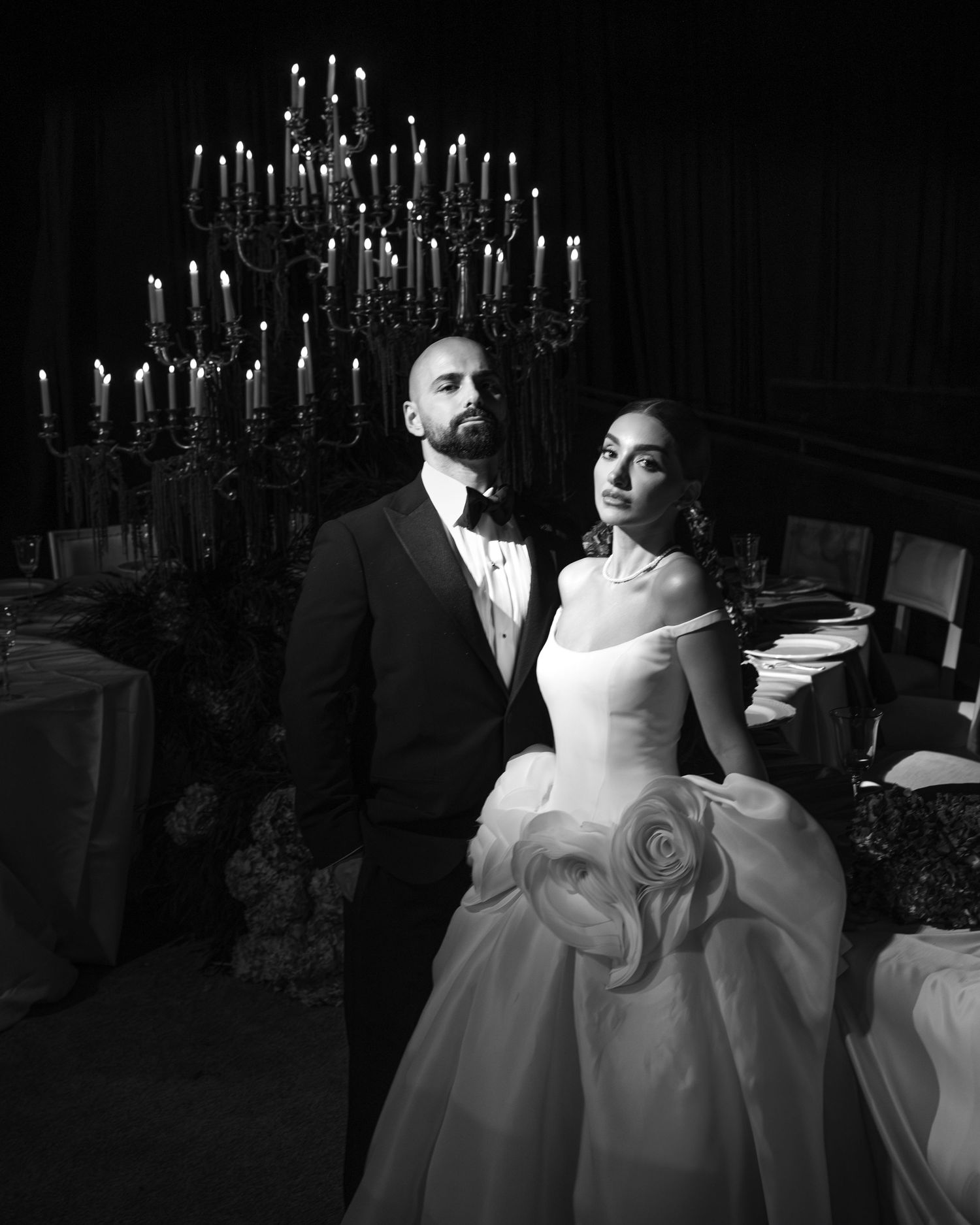 Black and white wedding portrait of bride in ball gown and groom in tuxedo beside candlelit candelabra.