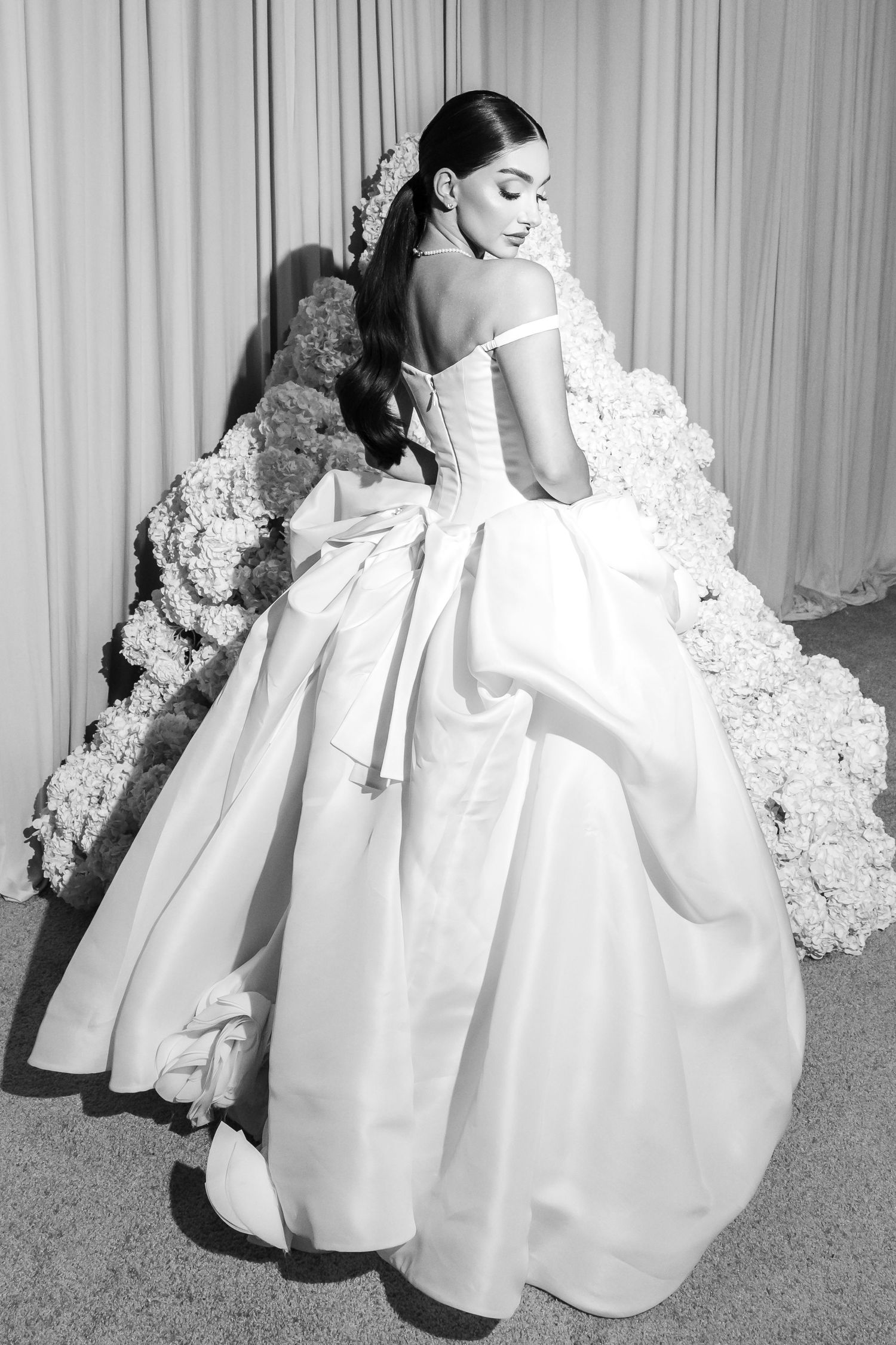 Black and white photo of a bride in an off-shoulder ball gown with bow back, posing near floral arrangements.