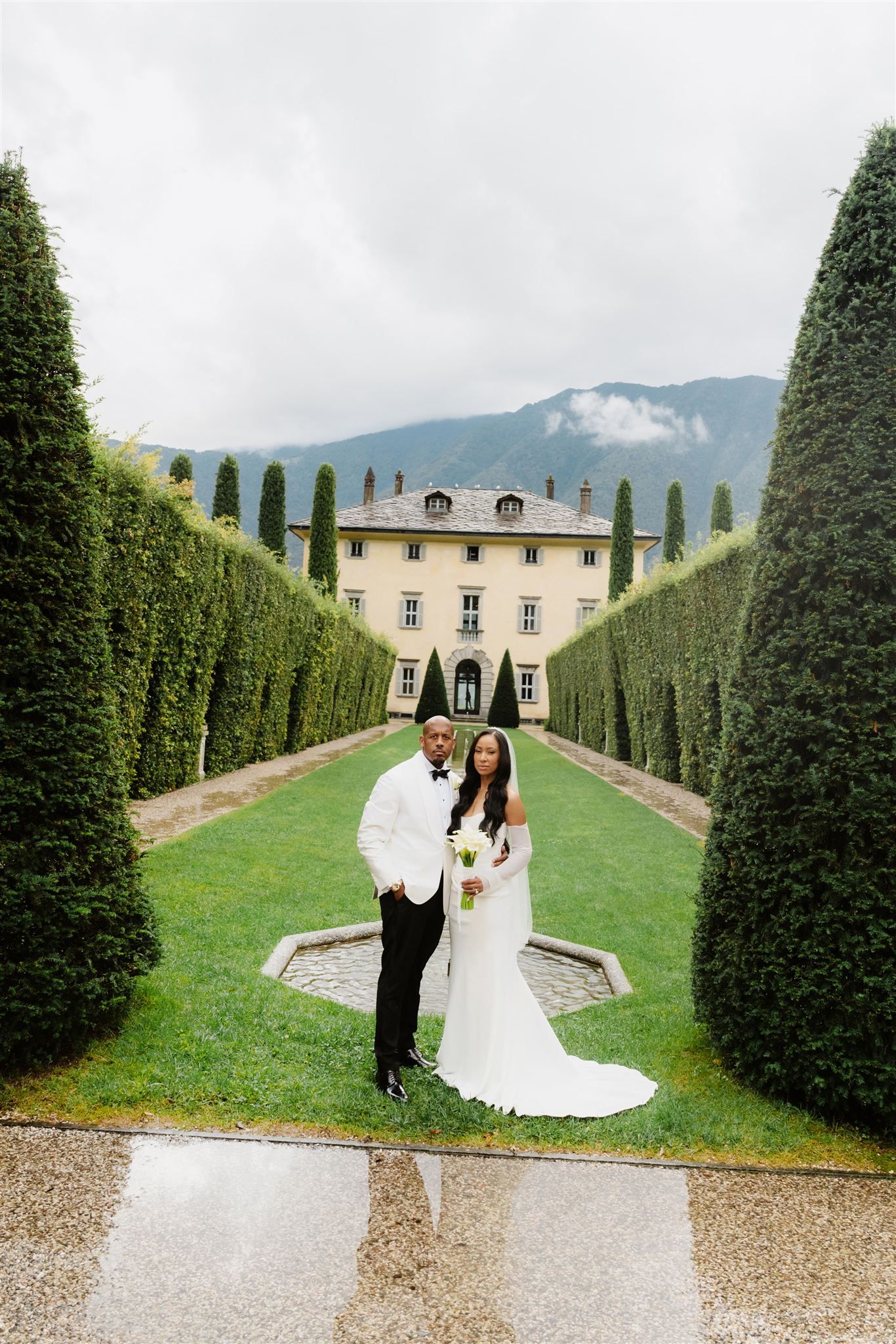 Wedding couple walking through a formal garden with cypress trees leading to an Italian villa.