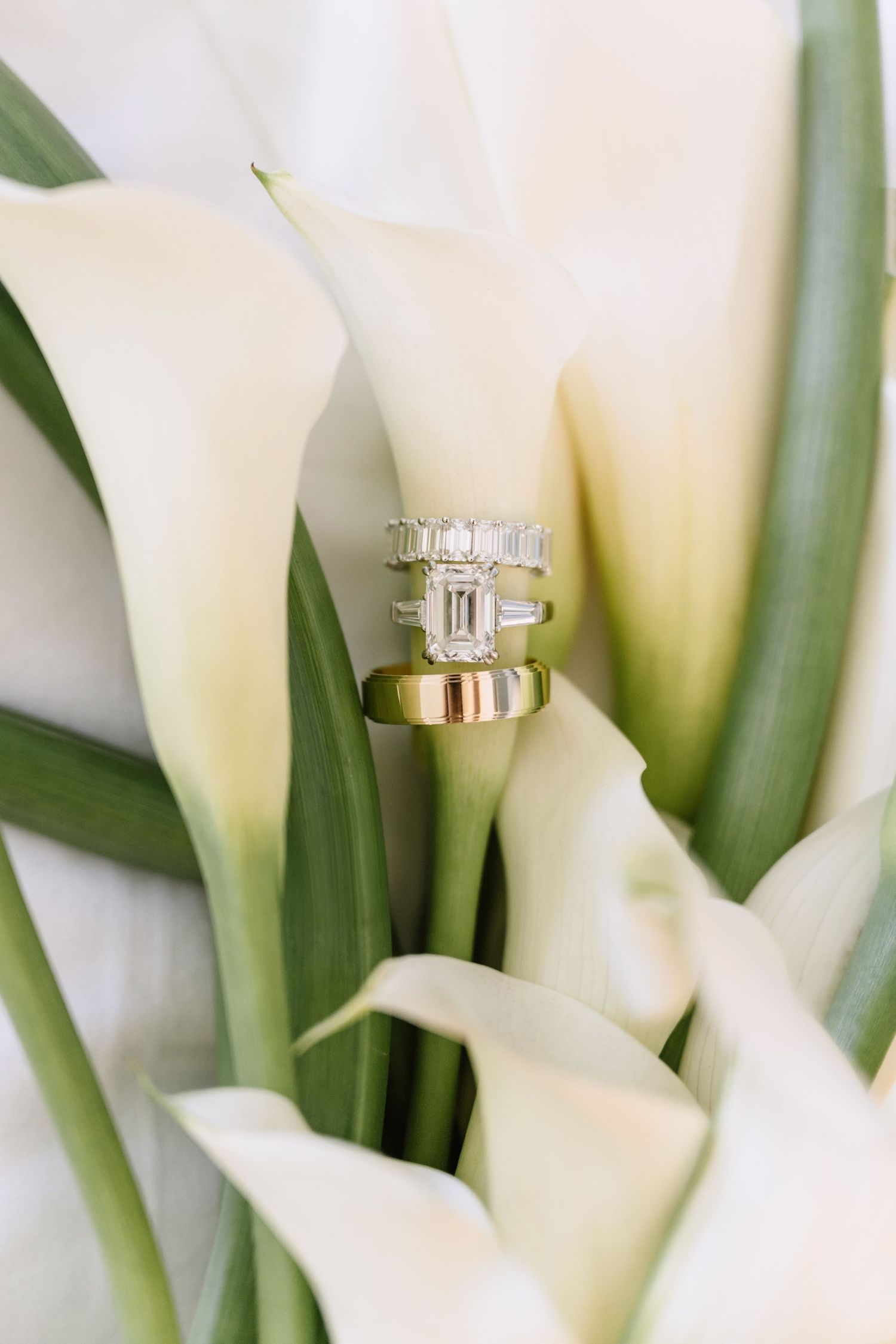 Elegant emerald cut diamond engagement ring with baguette wedding band and gold band nestled in white calla lilies.