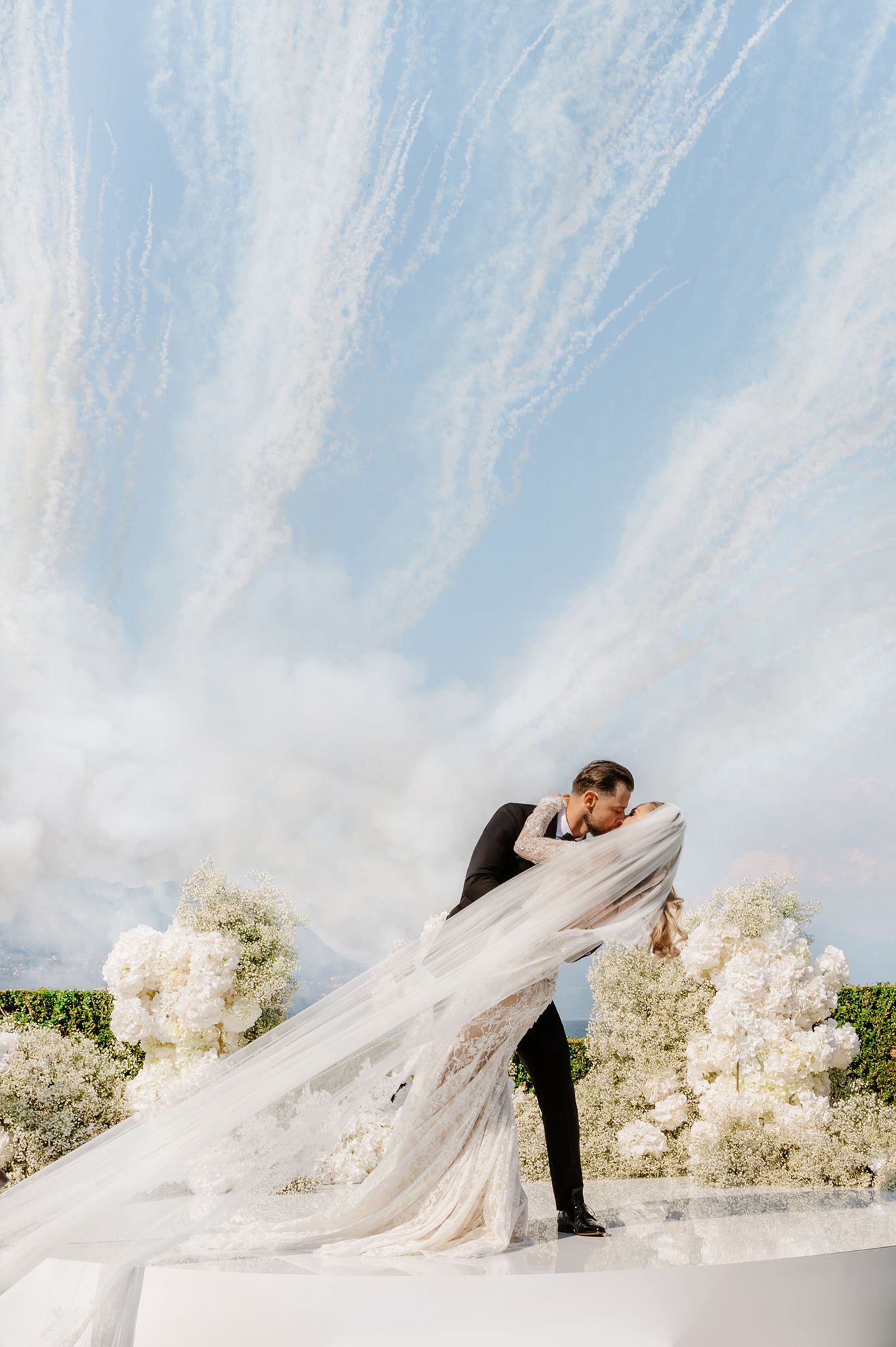 A flowing white wedding veil billows dramatically in the wind against a bright blue sky with white clouds.