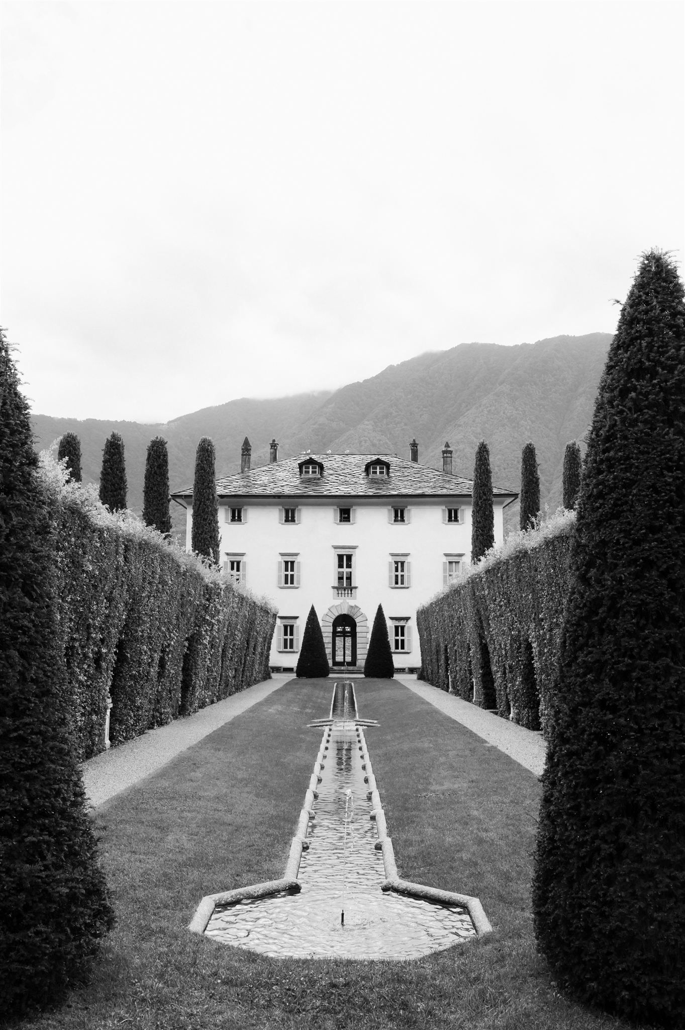 A grand white villa with symmetrical design and cypress trees lining the pathway up a hill.
