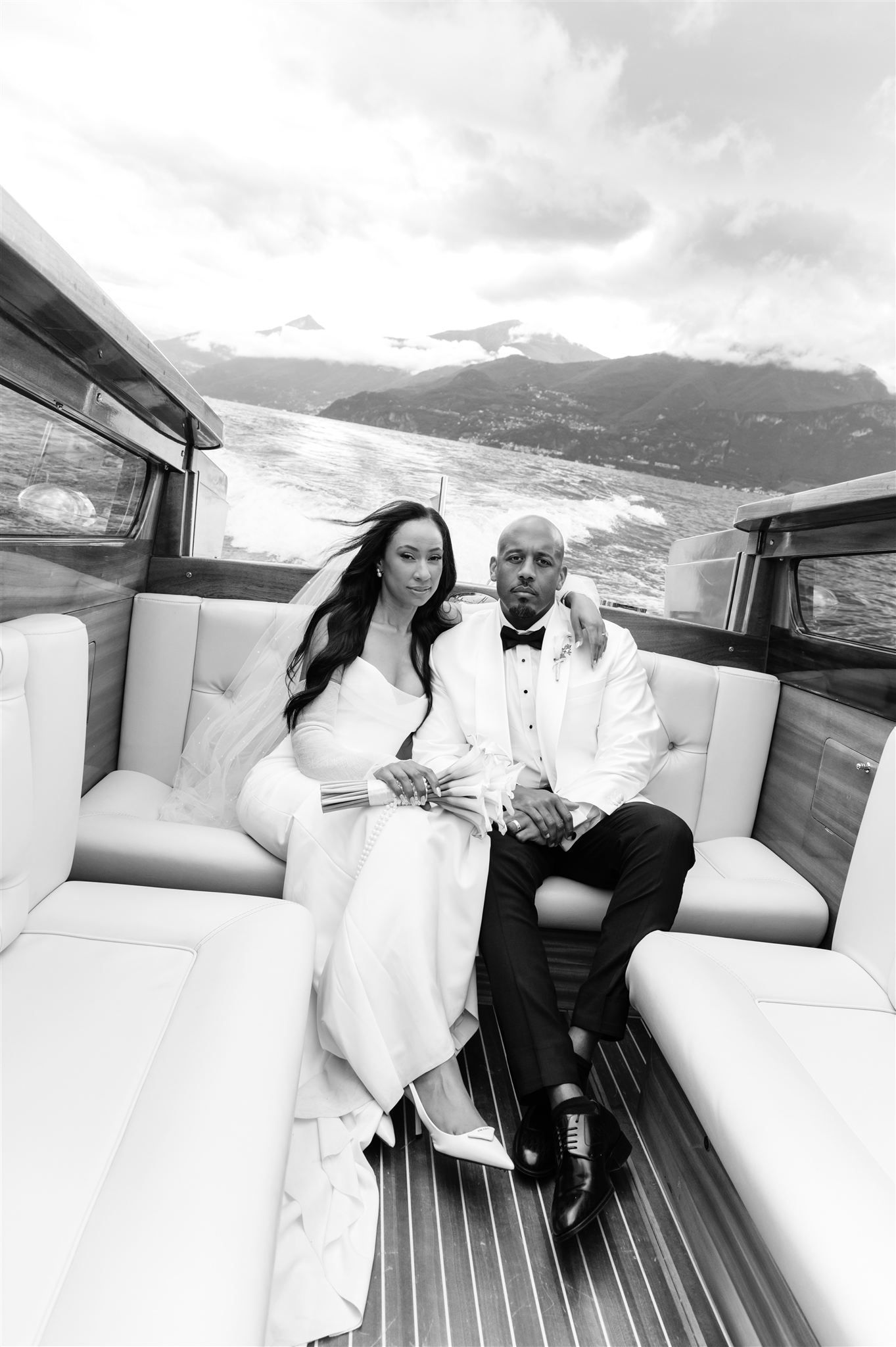 A couple relaxes on plush white seats of a luxury boat with mountain views in the background on Lake Como.