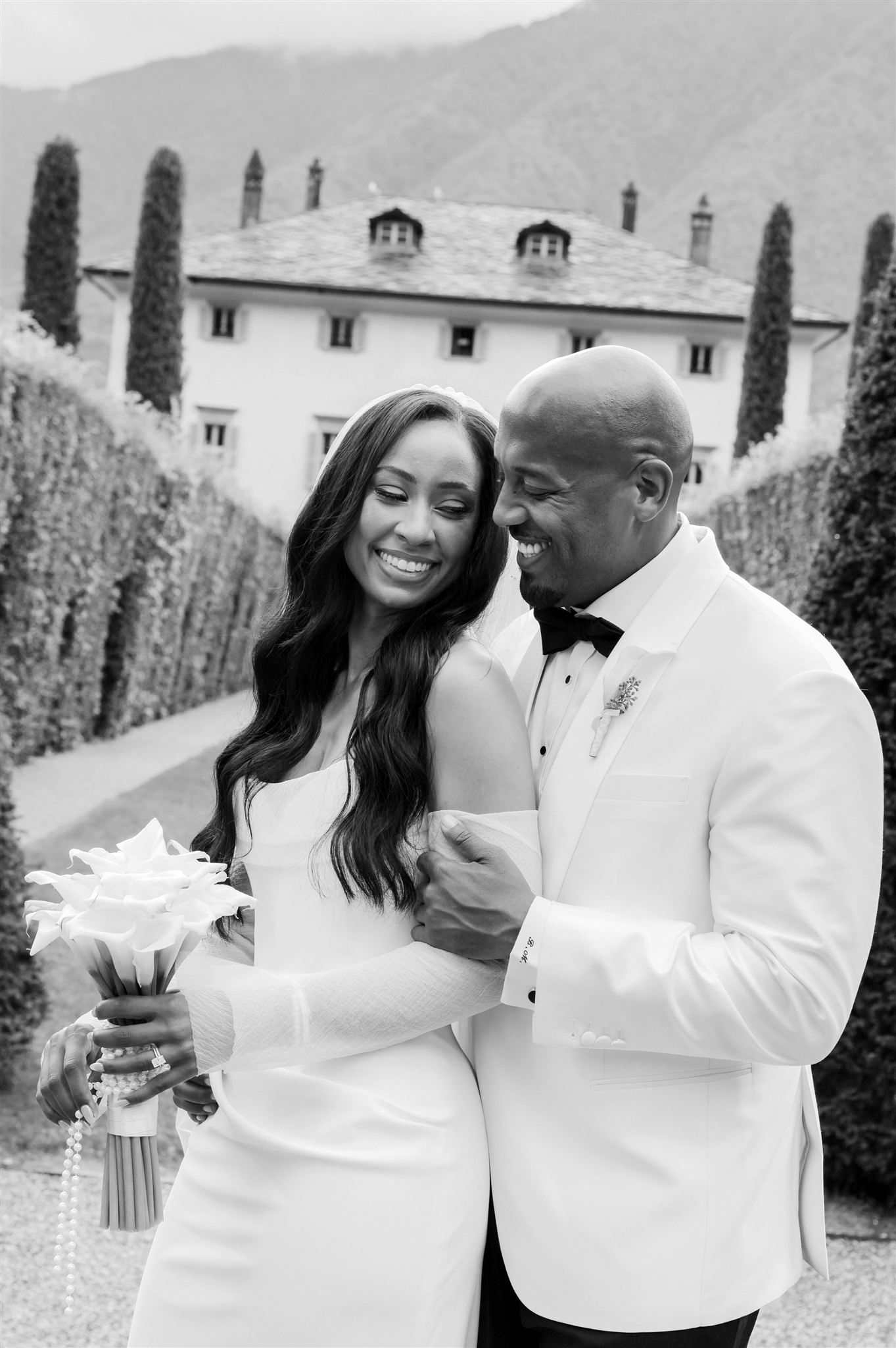A couple in matching white attire shares intimate moments against a mountain backdrop at Villa Balbiano