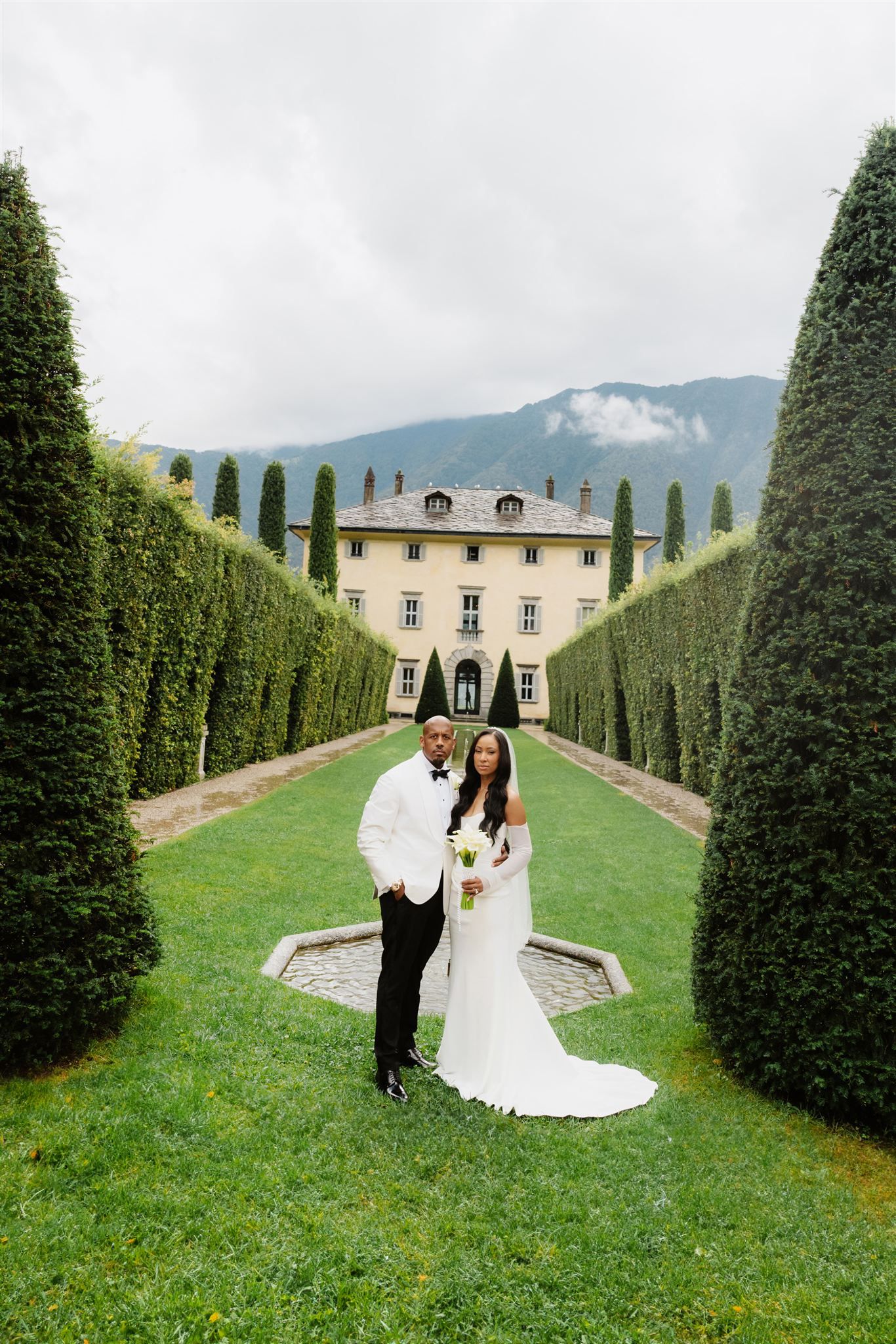 A couple in wedding attire walks through a manicured garden path leading to a grand Italian Villa Balbiano.