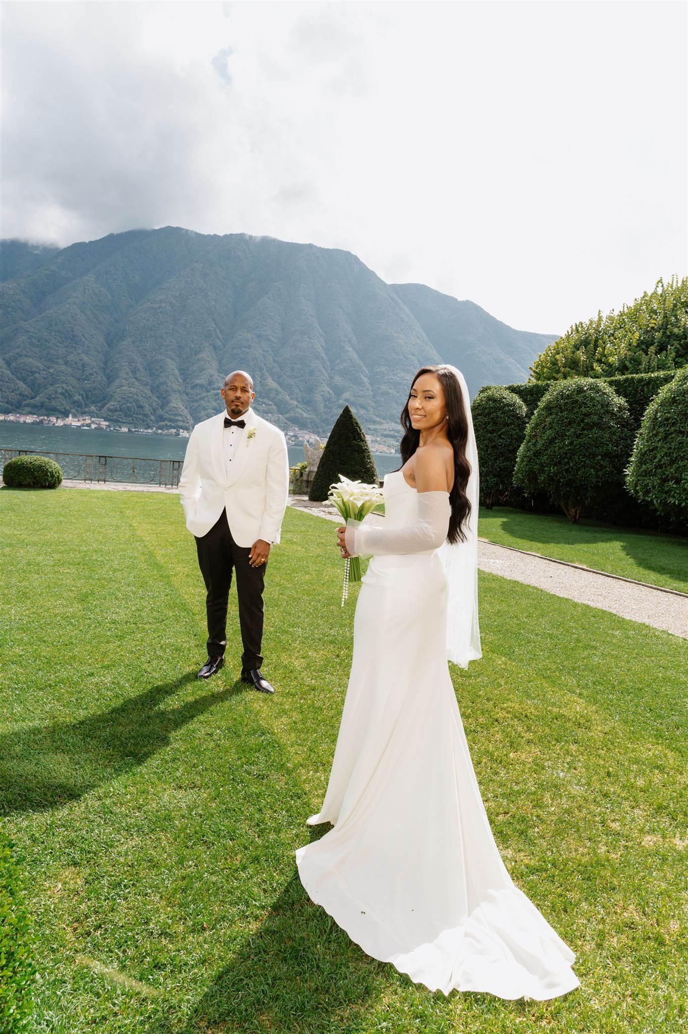 Elegant wedding couple poses in a lush garden with mountain views and manicured hedges in the background.