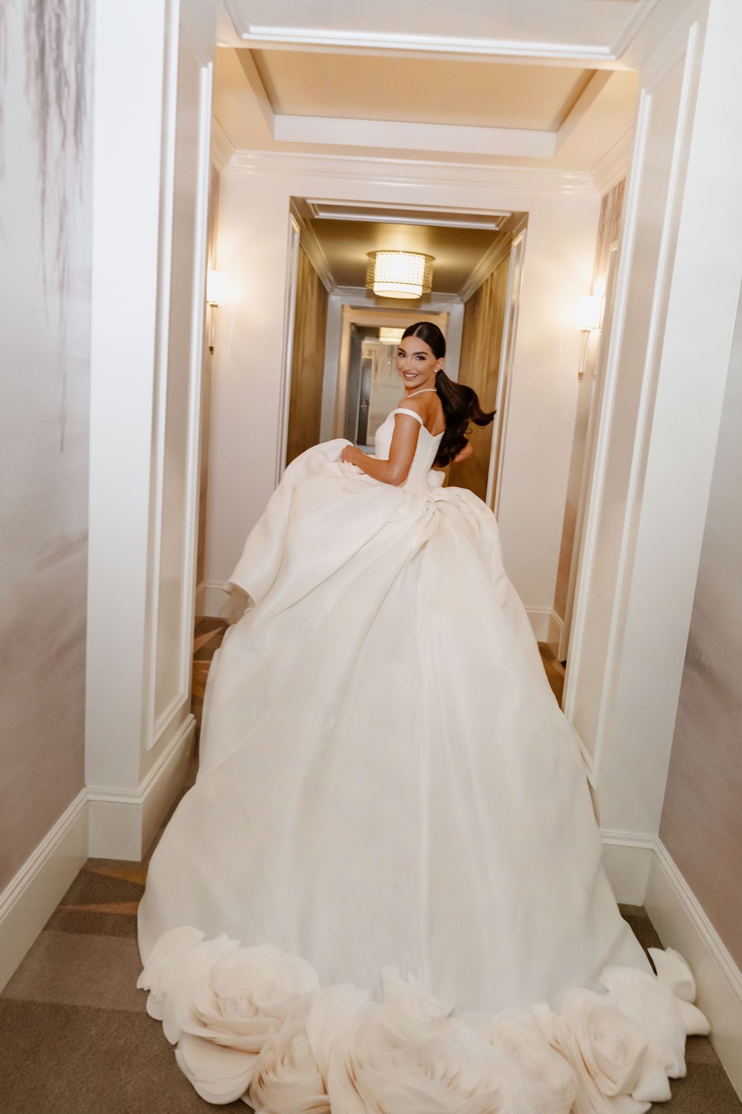 Bride in a voluminous white ball gown with floral hem detail smiling in an elegant hotel corridor.