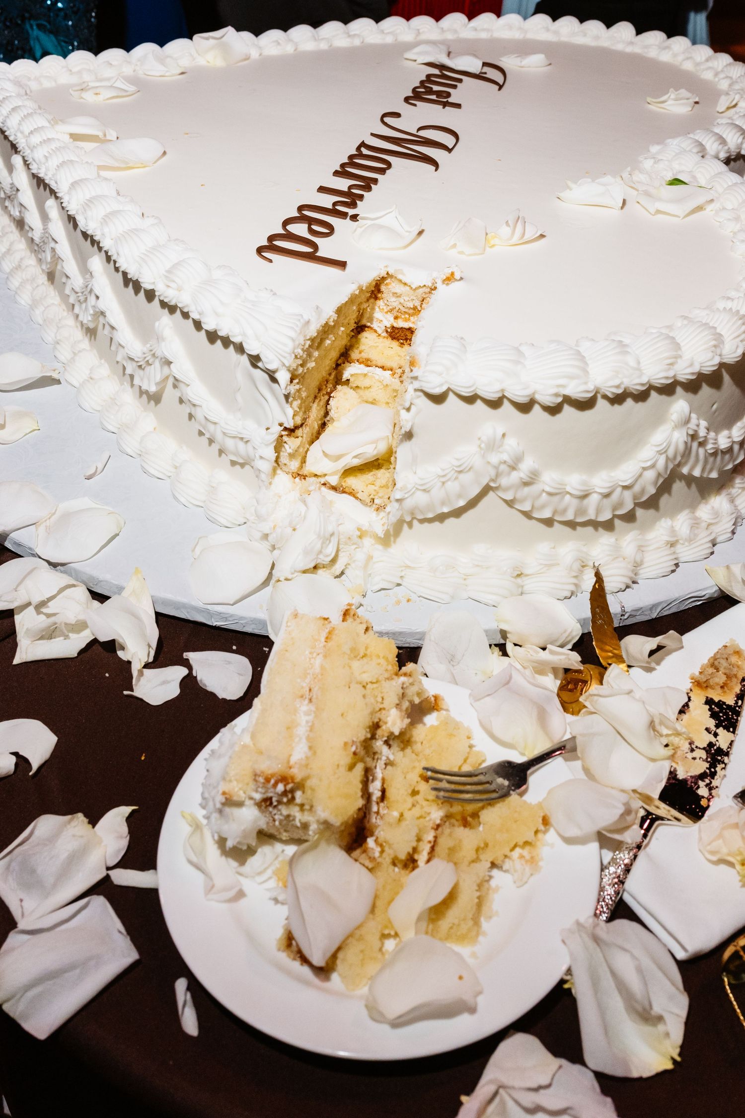 A just married wedding cake with a slice cut out, served on a plate with a fork, surrounded by white rose petals.