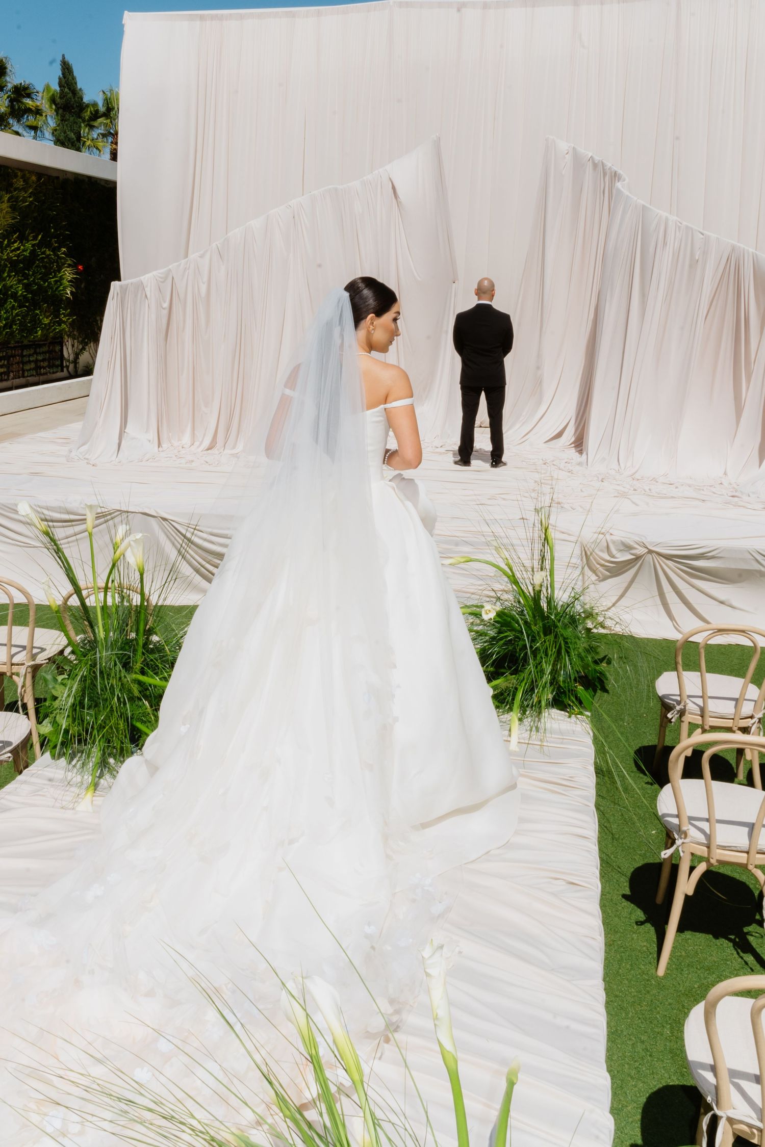 Bride in off-shoulder ball gown with long veil walks toward groom at outdoor wedding ceremony with white drape backdrop.