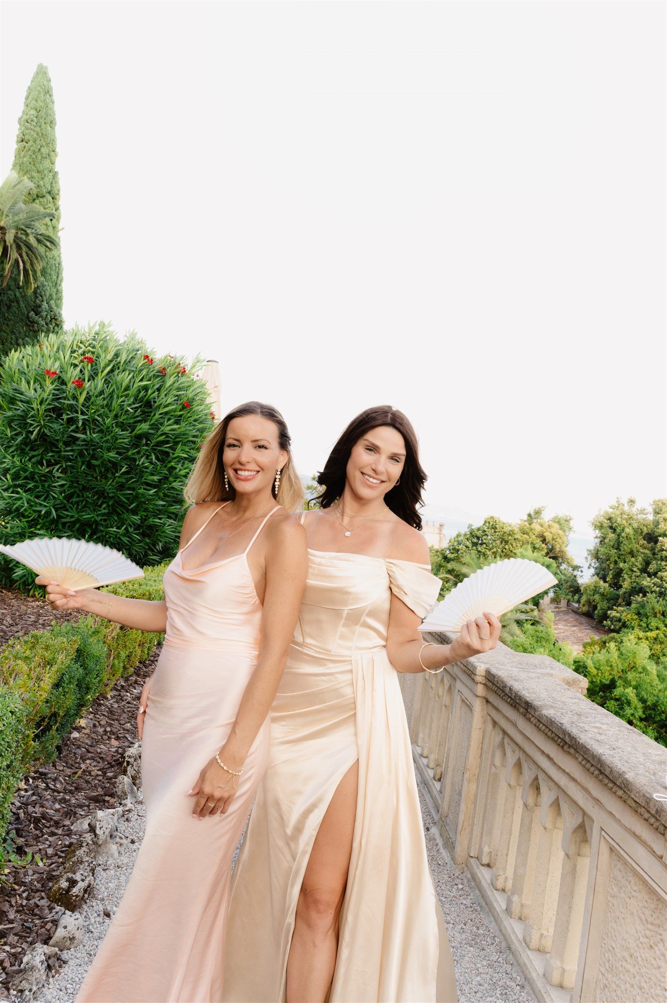 Two bridesmaids in elegant champagne-colored dresses pose on a stone balcony surrounded by lush greenery.