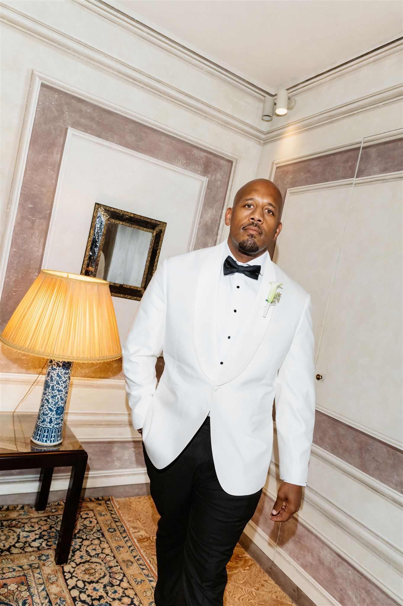 Person in formal white tuxedo jacket and bowtie posing in elegant room with table lamp.